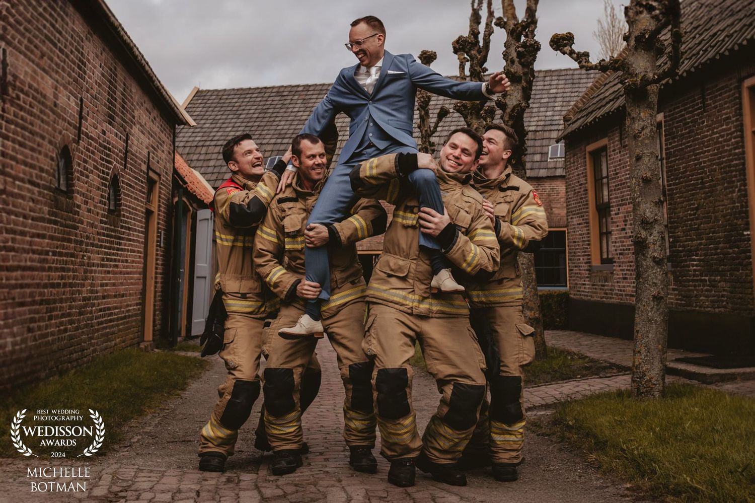 Group of firefighters in tan uniforms working together to lift colleague in blue in alleyway between brick buildings.