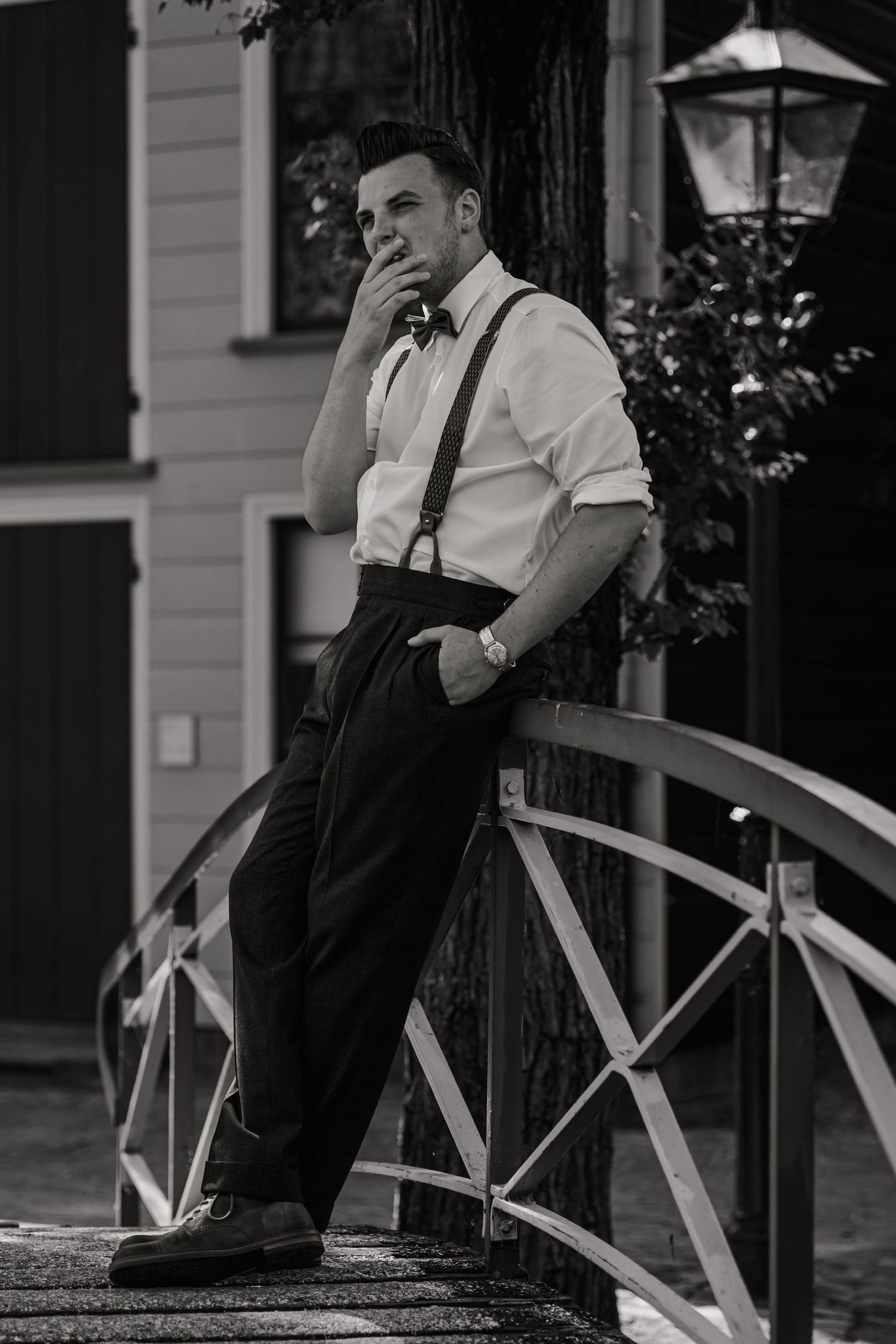 Groom in formal attire sits thoughtfully on outdoor porch railing.