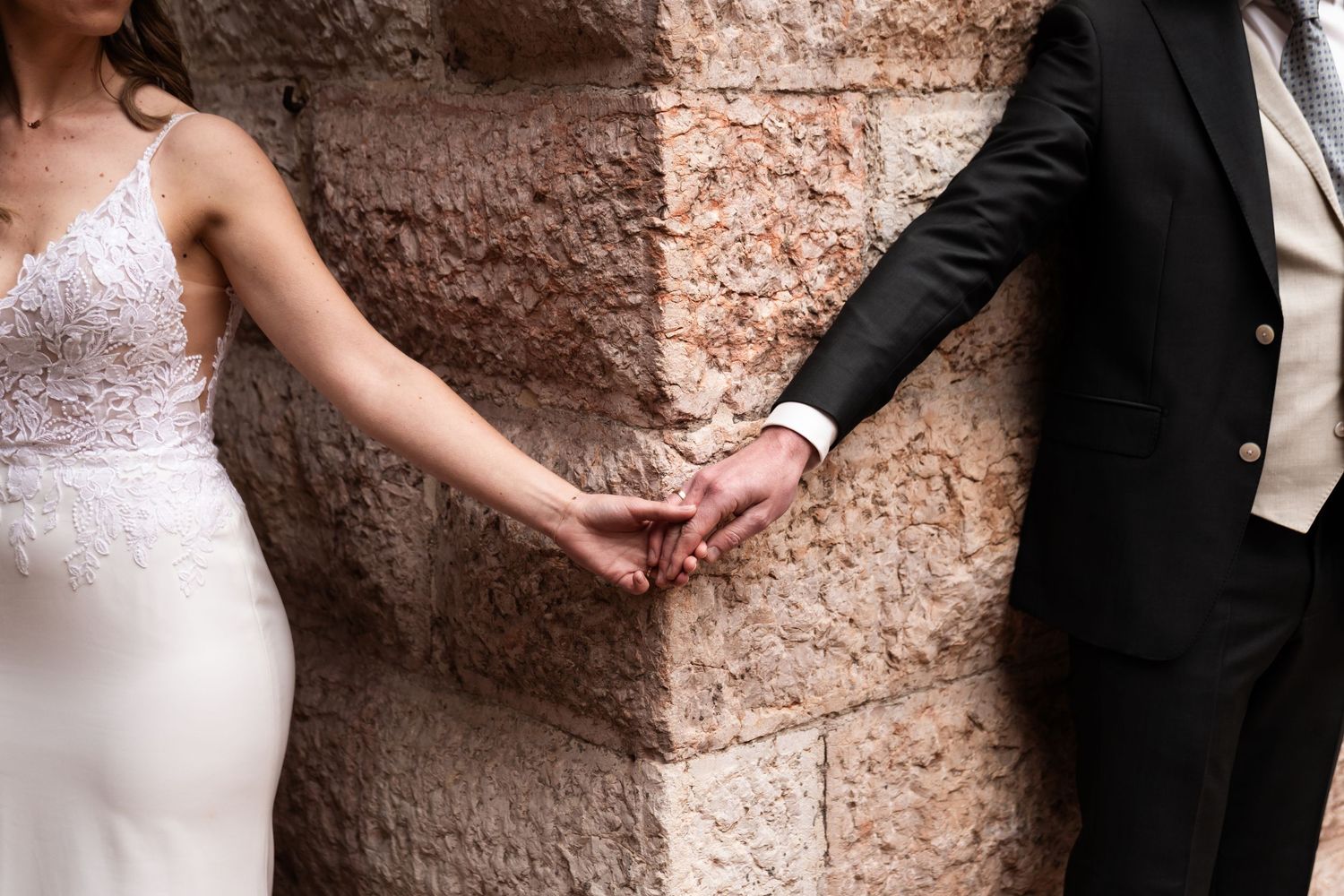 A bride and groom hold hands against a rustic stone wall background in a romantic wedding moment.