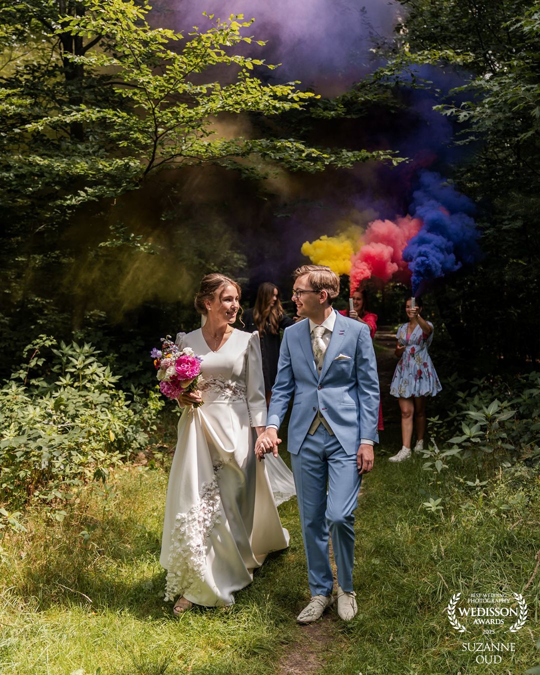 A couple walks through a wooded path with colorful smoke bombs creating vibrant pink and blue hues in nature.