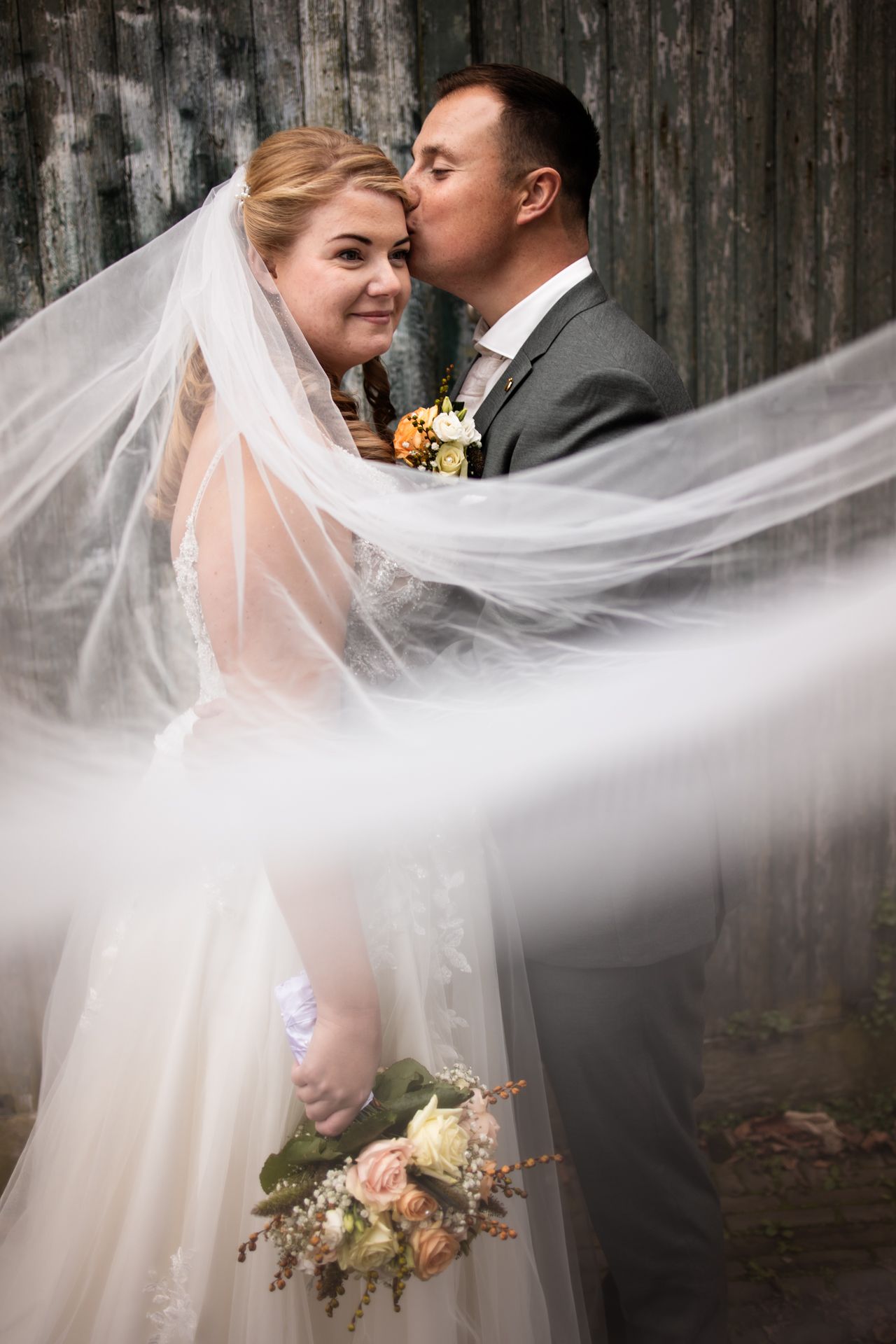 A romantic wedding moment captured with flowing veil and rustic wood background as newlyweds share a tender embrace.