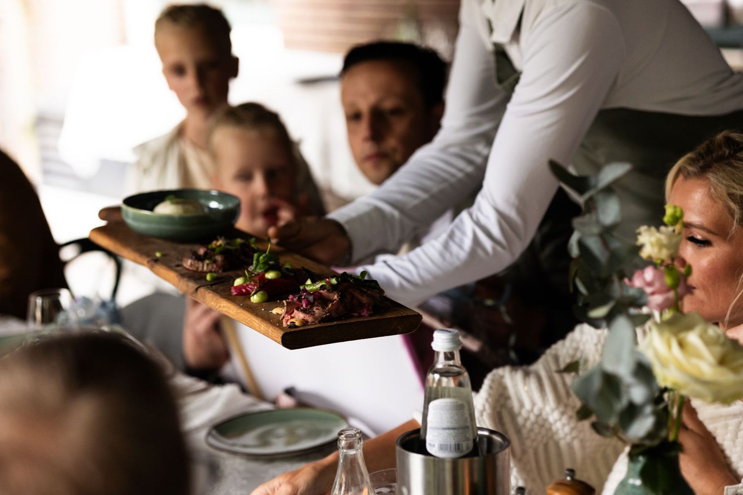Server presenting a large platter of food to guests at an elegant dining table with floral arrangements.