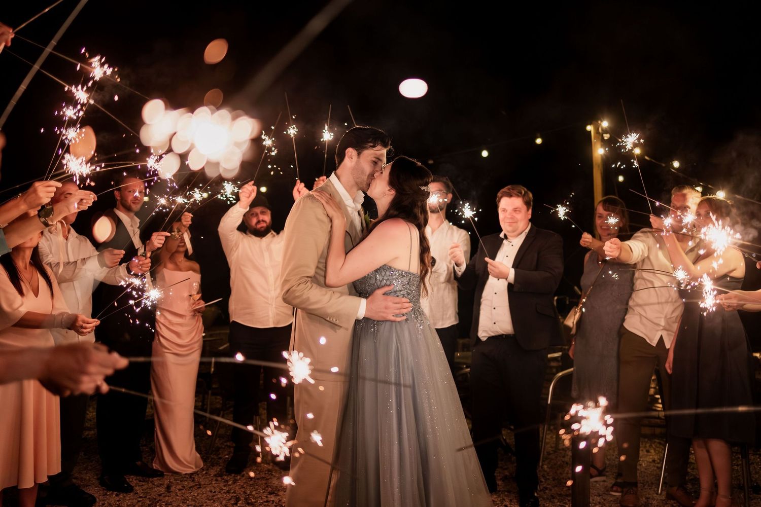 Wedding guests hold sparklers creating a magical nighttime tunnel for the newlyweds at an outdoor celebration.