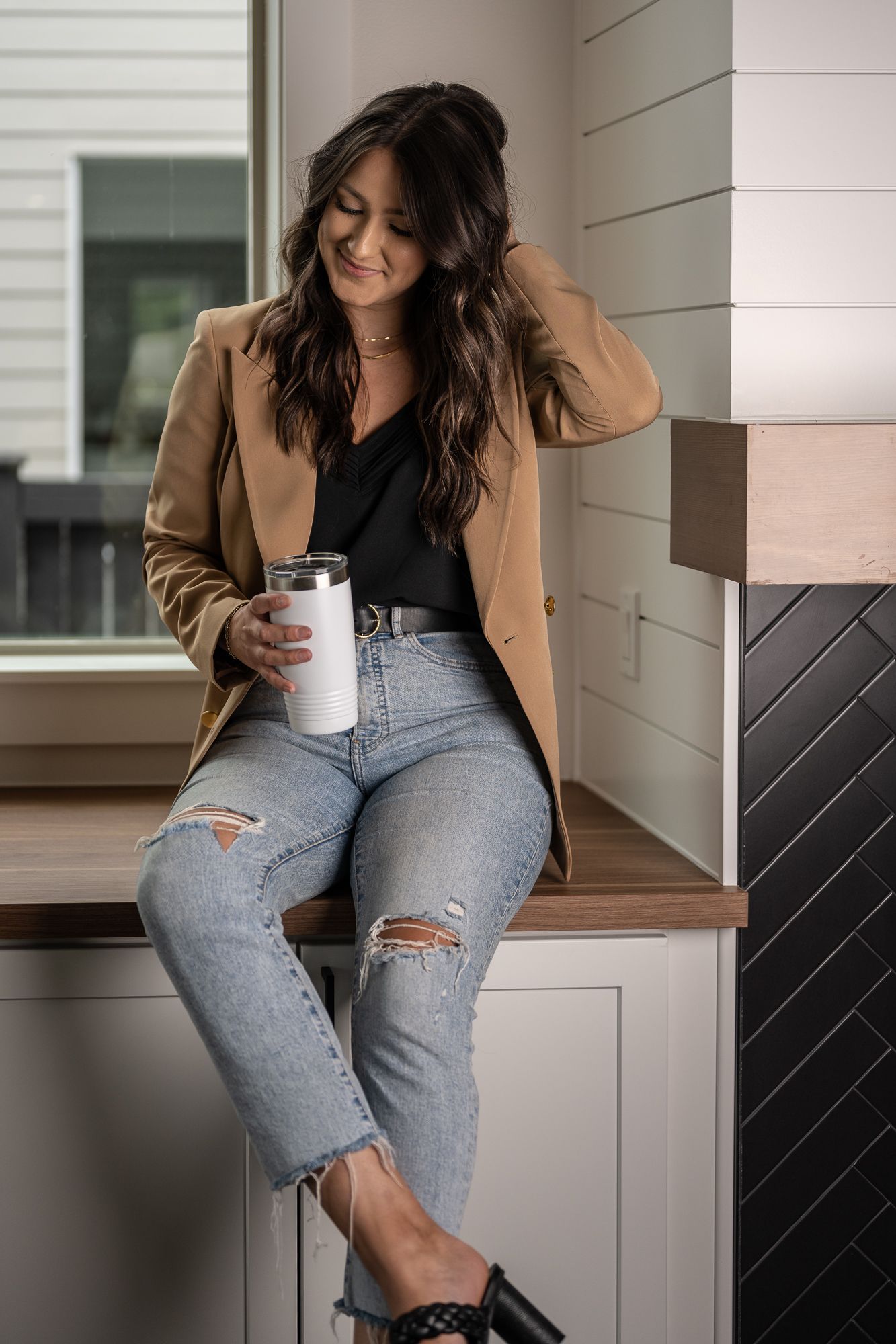 Casual portrait of person sitting on counter wearing camel blazer, ripped jeans and holding white tumbler.