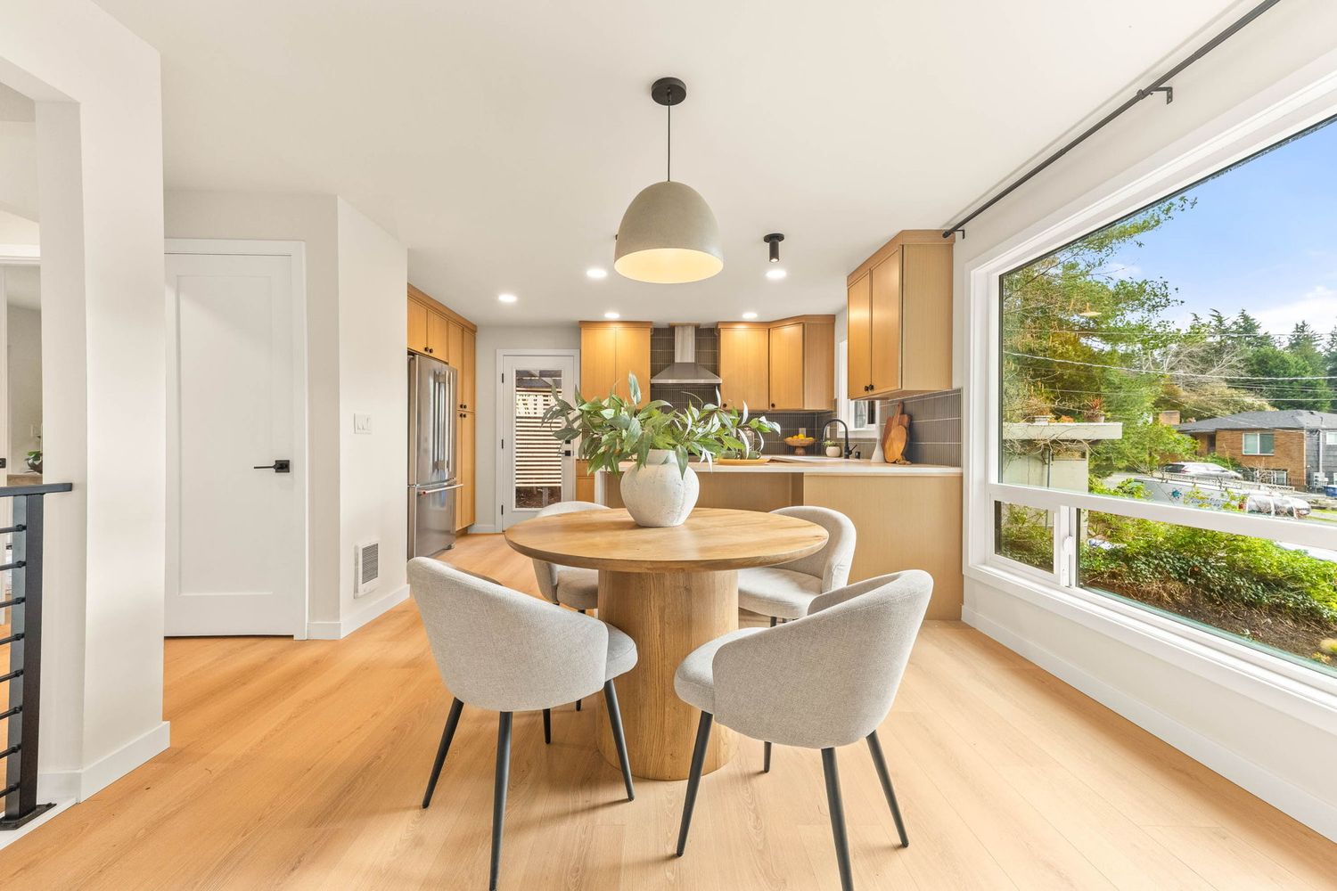 Bright dining area with round wooden table, pendant light, and large window overlooking outdoor scenery.