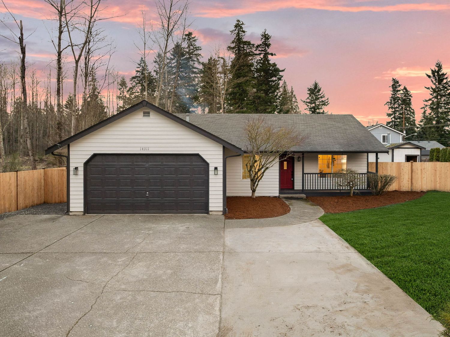 A concrete driveway leads to a residential garage under a pink and orange sunset sky with bare trees in winter.