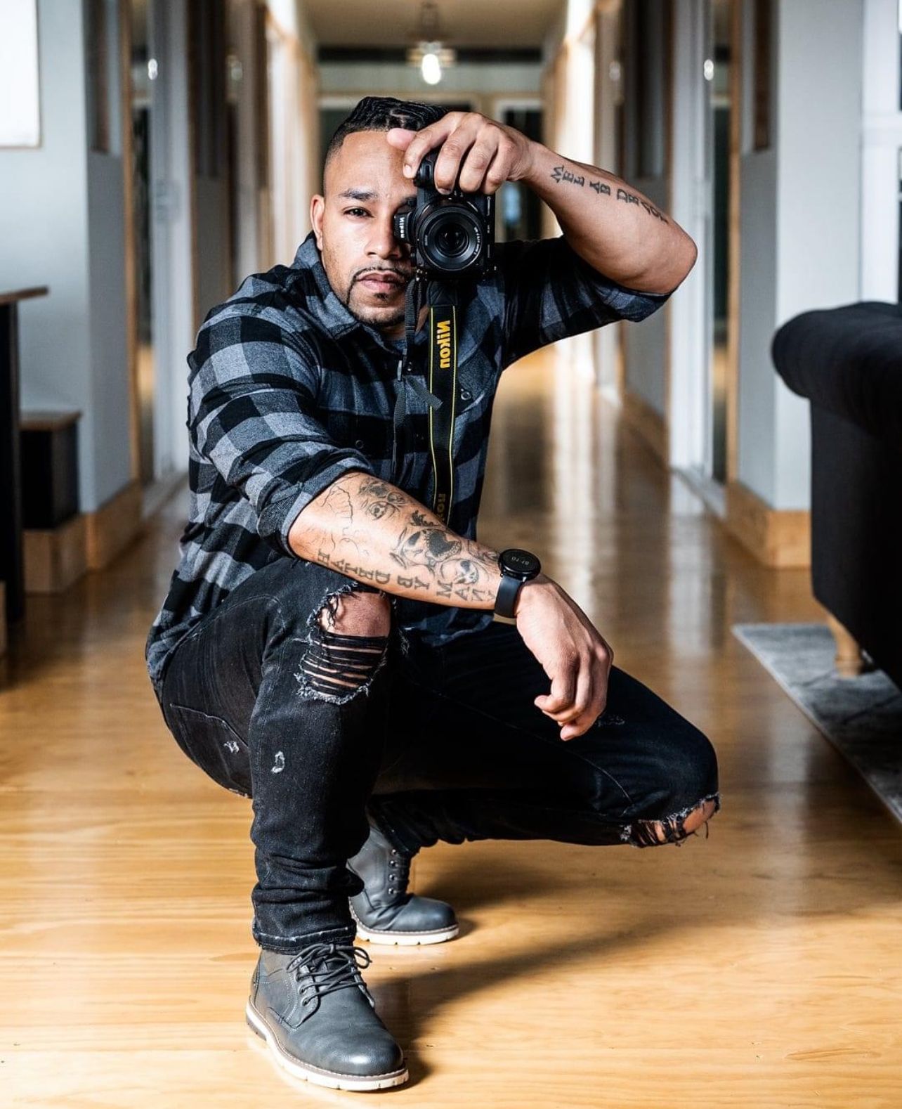 A photographer in plaid shirt and ripped jeans crouches in a hallway holding a professional camera.