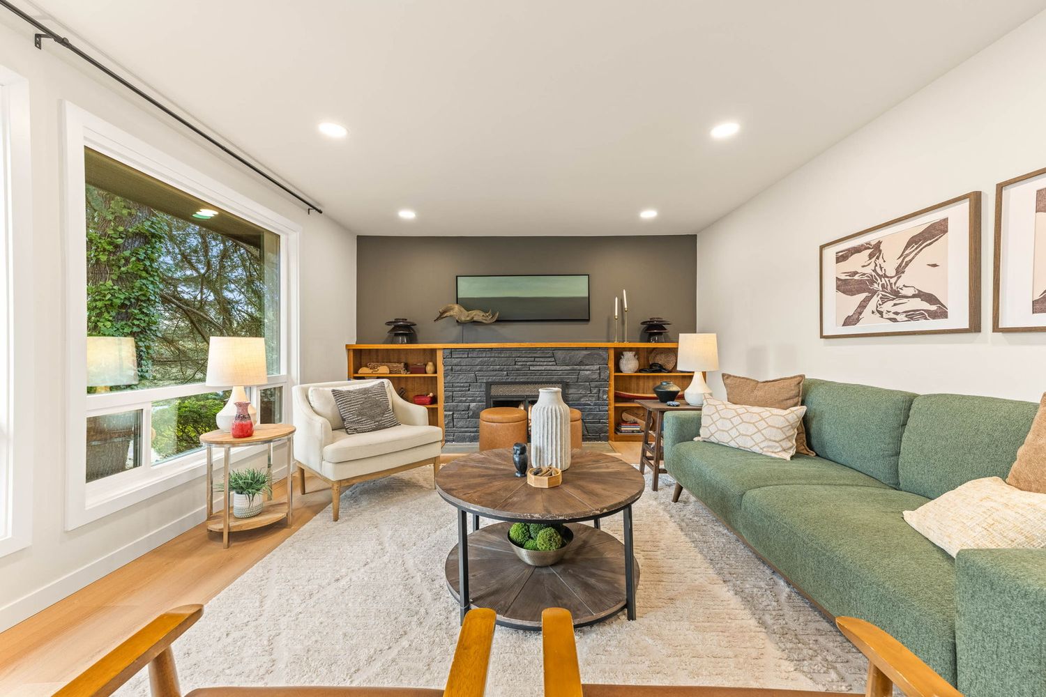 Modern living room features a green sofa, round coffee table, and fireplace with natural light from large windows.