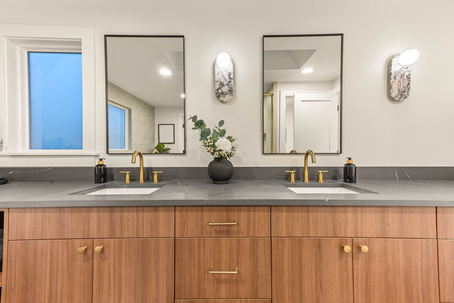 Modern bathroom vanity with wood cabinets, gray countertop, dual mirrors, brass faucets and wall sconces.