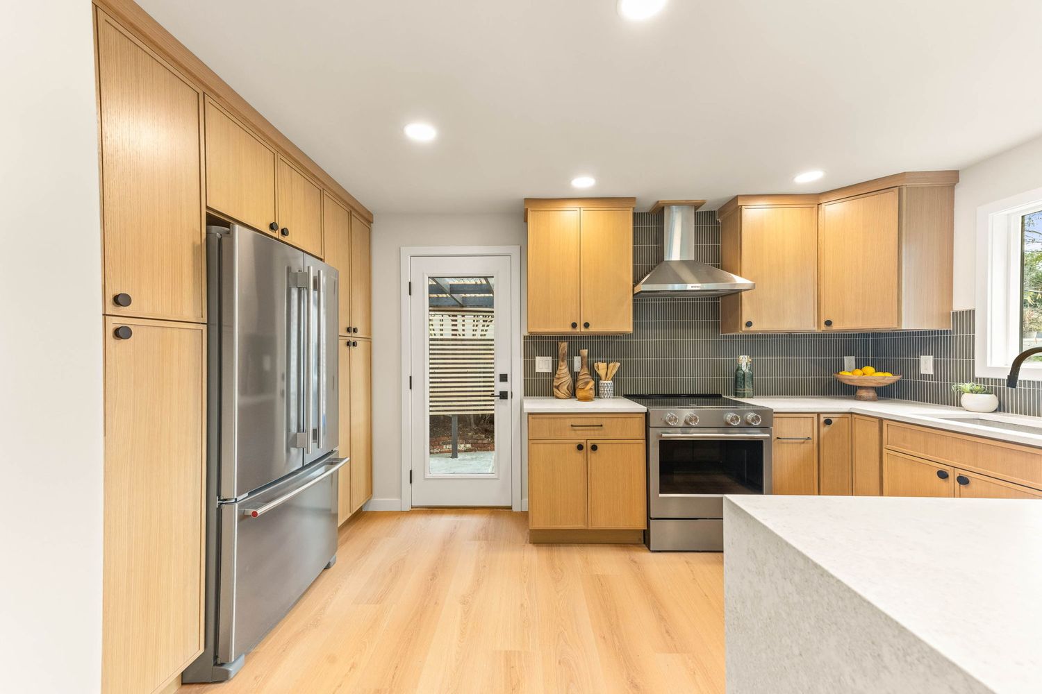 Modern kitchen with light wood cabinets, stainless steel appliances, gray backsplash, and hardwood flooring.