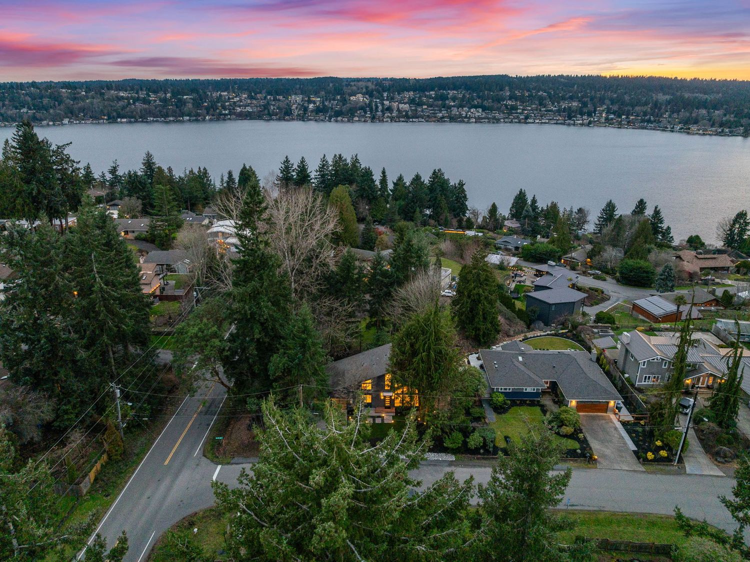 Aerial view of lakefront homes and winding roads nestled among evergreen trees at sunset with pink and purple skies.