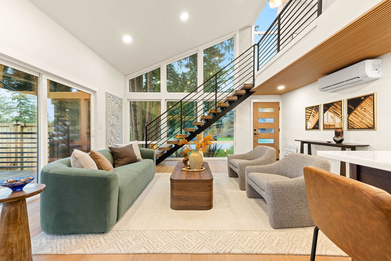 Modern living room with high ceilings, floating staircase, green sofa, and large windows overlooking trees.