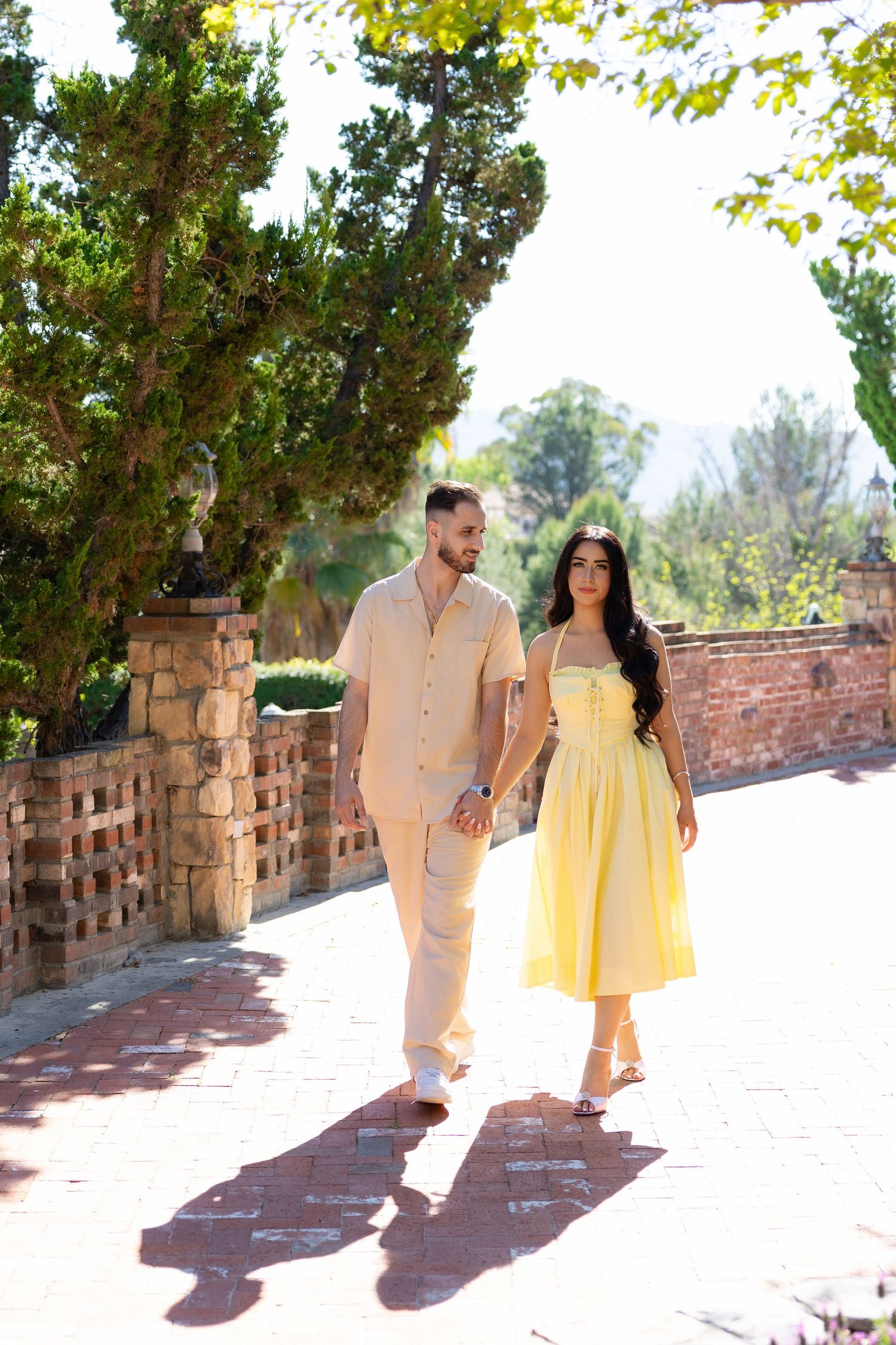 Couple holding hands walking on brick path in garden, man in beige outfit and woman in yellow sundress.