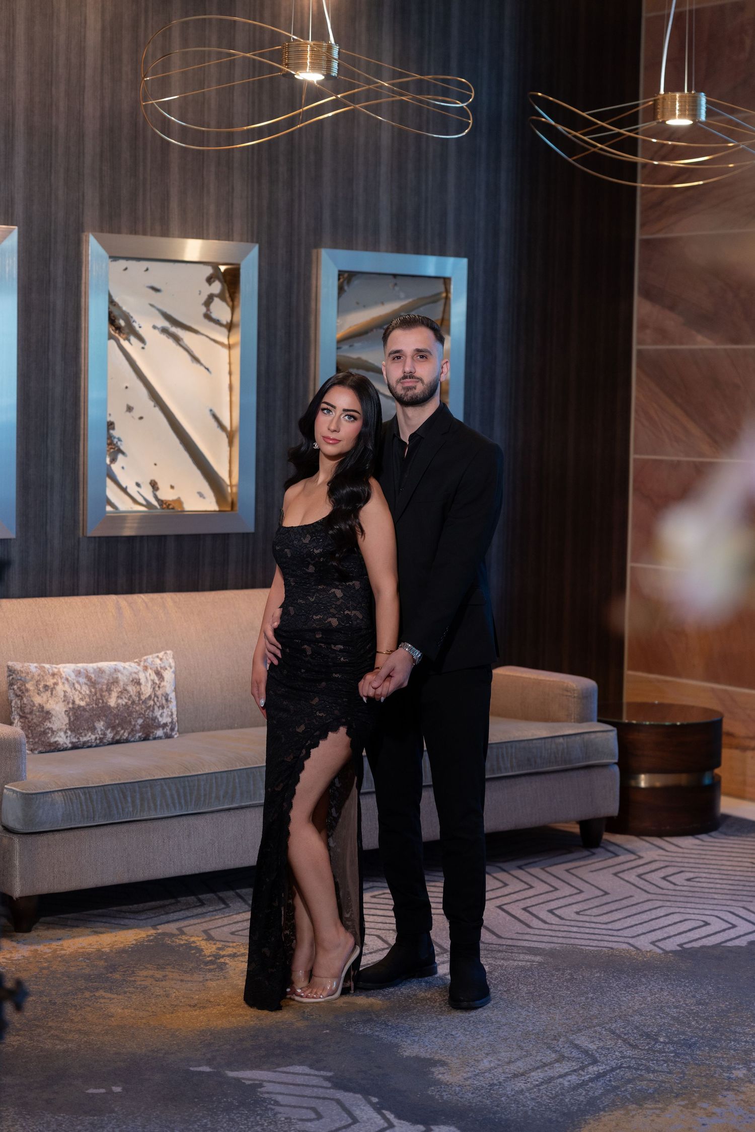 Elegant couple in matching black formal attire posing in a luxurious hotel lobby with modern art and gold chandeliers.