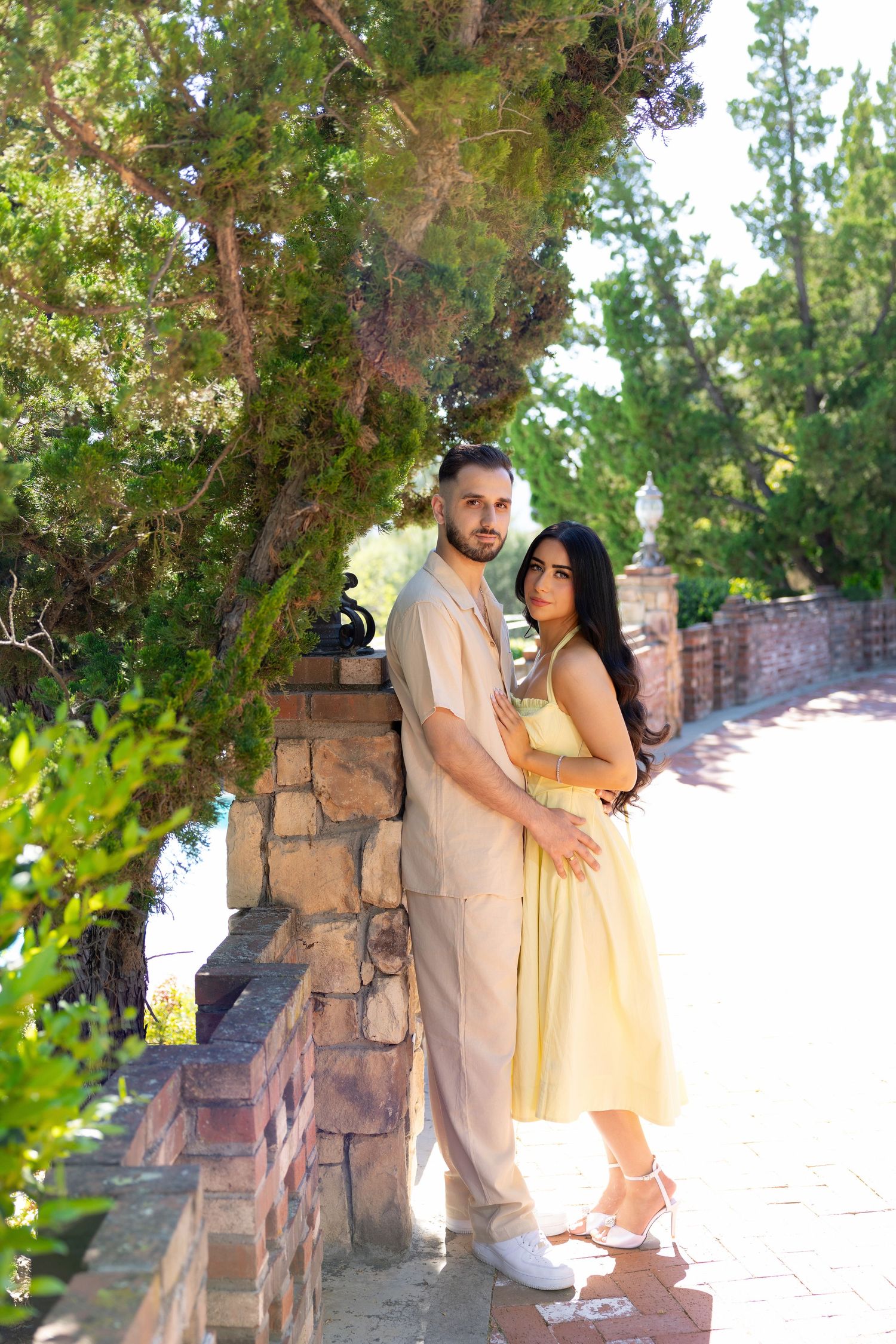 Couple in matching beige outfits posing in a lush garden with stone walls and green trees.