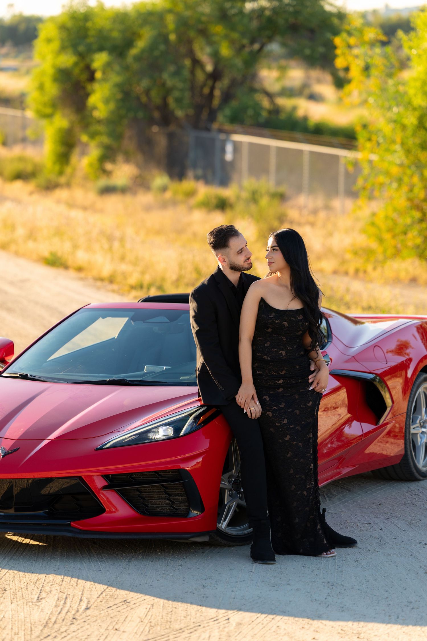 Couple in black outfits posing by a red Corvette sports car on a dirt road at golden hour.
