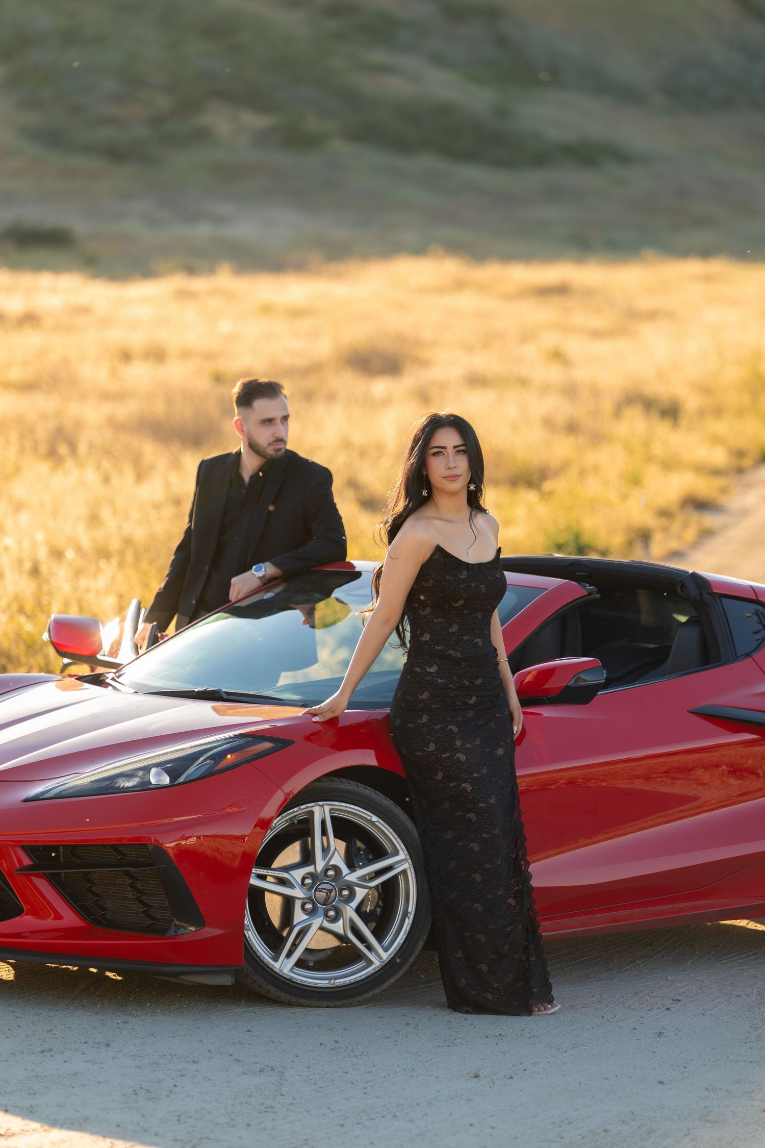 Couple in elegant black formalwear posing with a red Corvette sports car in a scenic golden field at sunset.