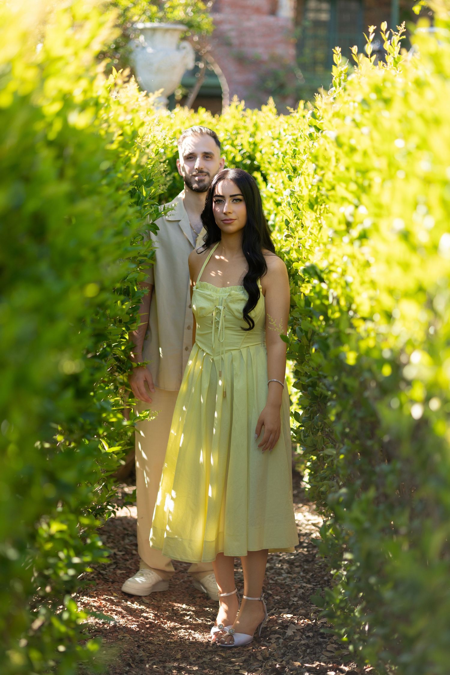 Couple posing in a sunlit garden path, woman in yellow midi dress, man in beige coat, surrounded by lush green hedges.
