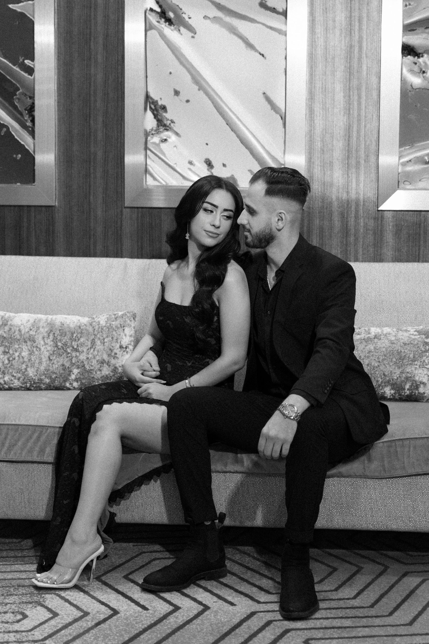 Black and white photo of an elegant couple in formal attire sitting closely on a luxurious sofa.