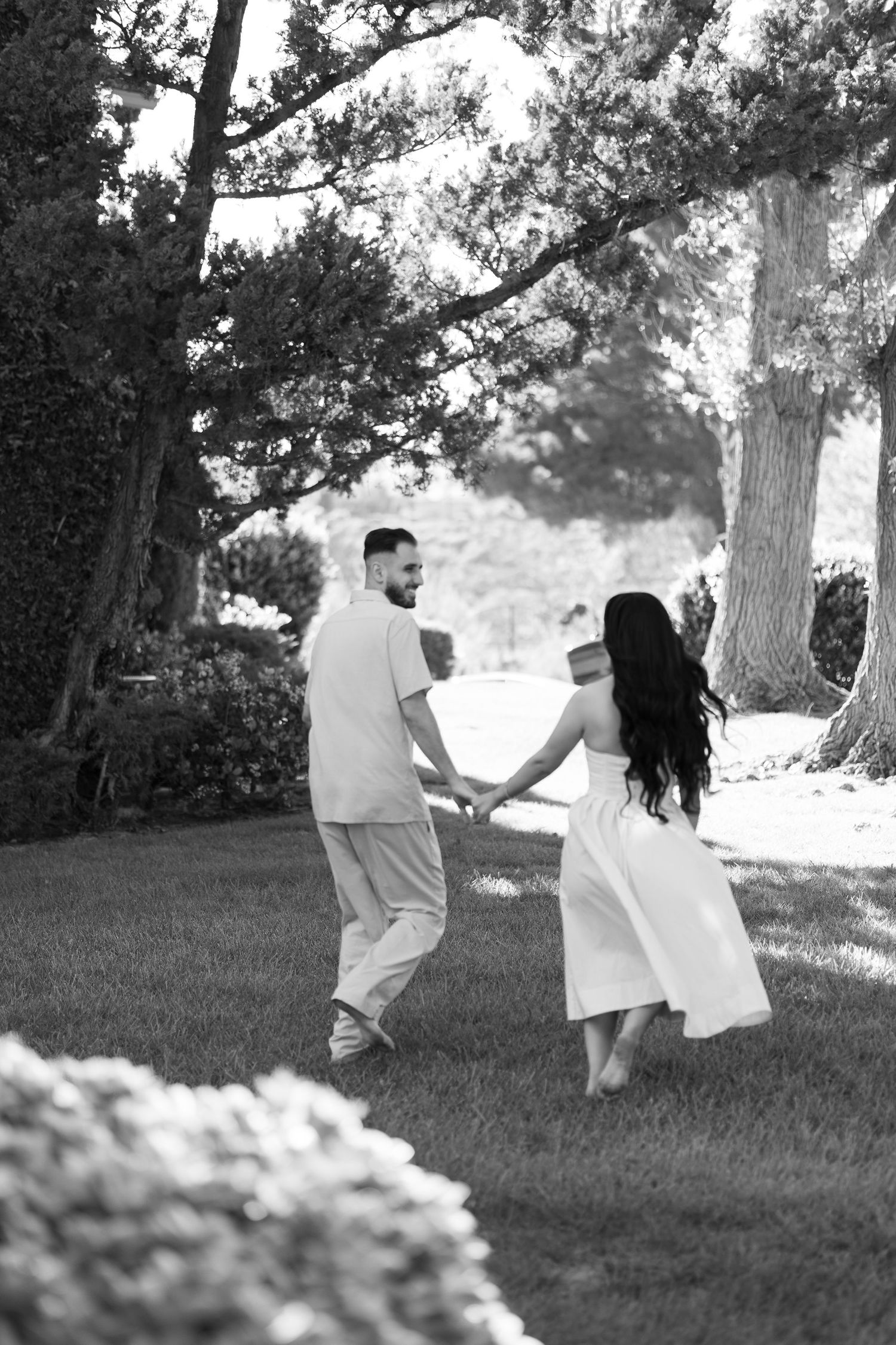 Black and white photo of a couple holding hands and walking through a lush green park, woman in white dress.