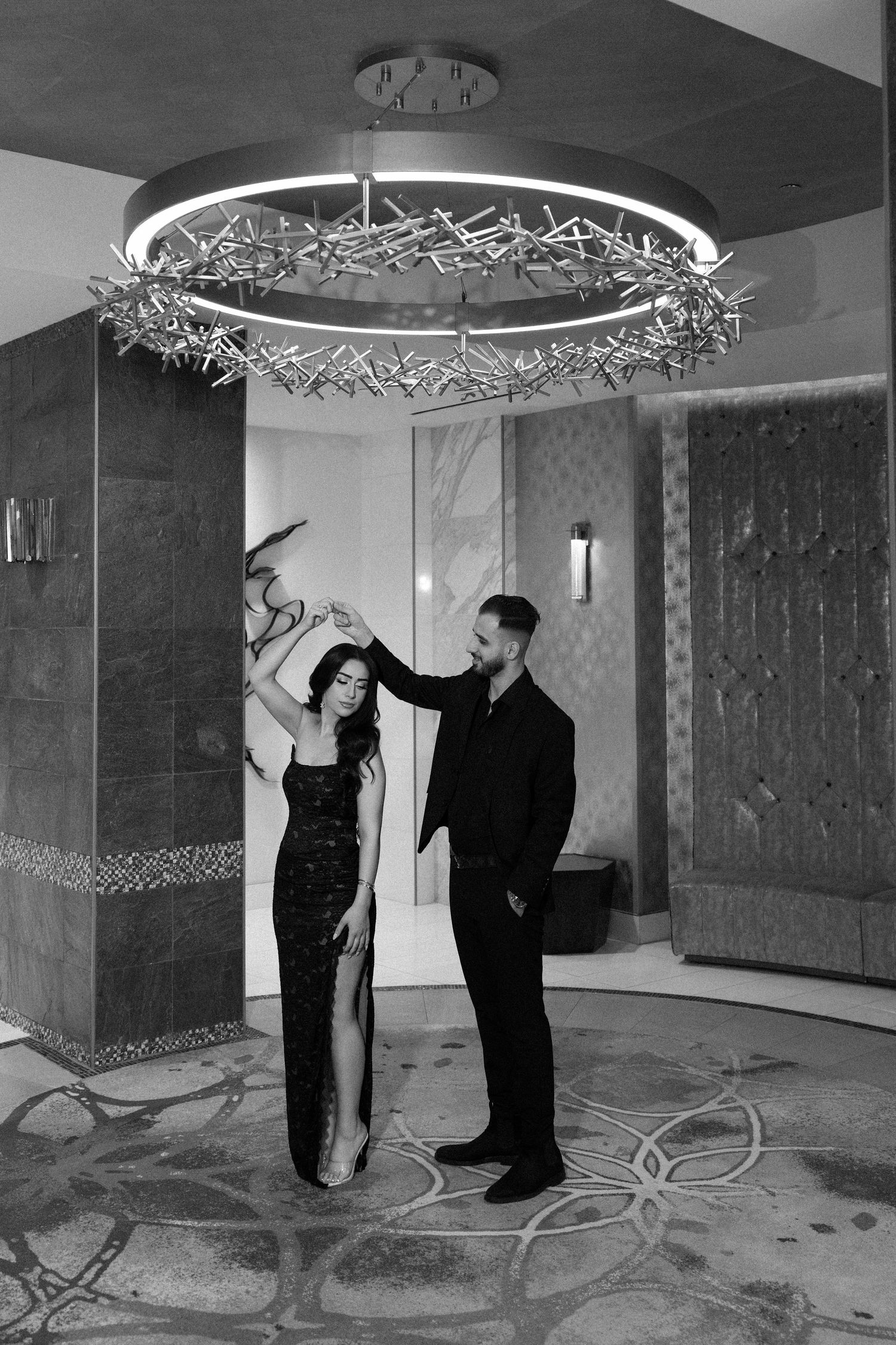 Black and white photo of an elegant couple dancing under a decorative chandelier in a luxurious hotel lobby.
