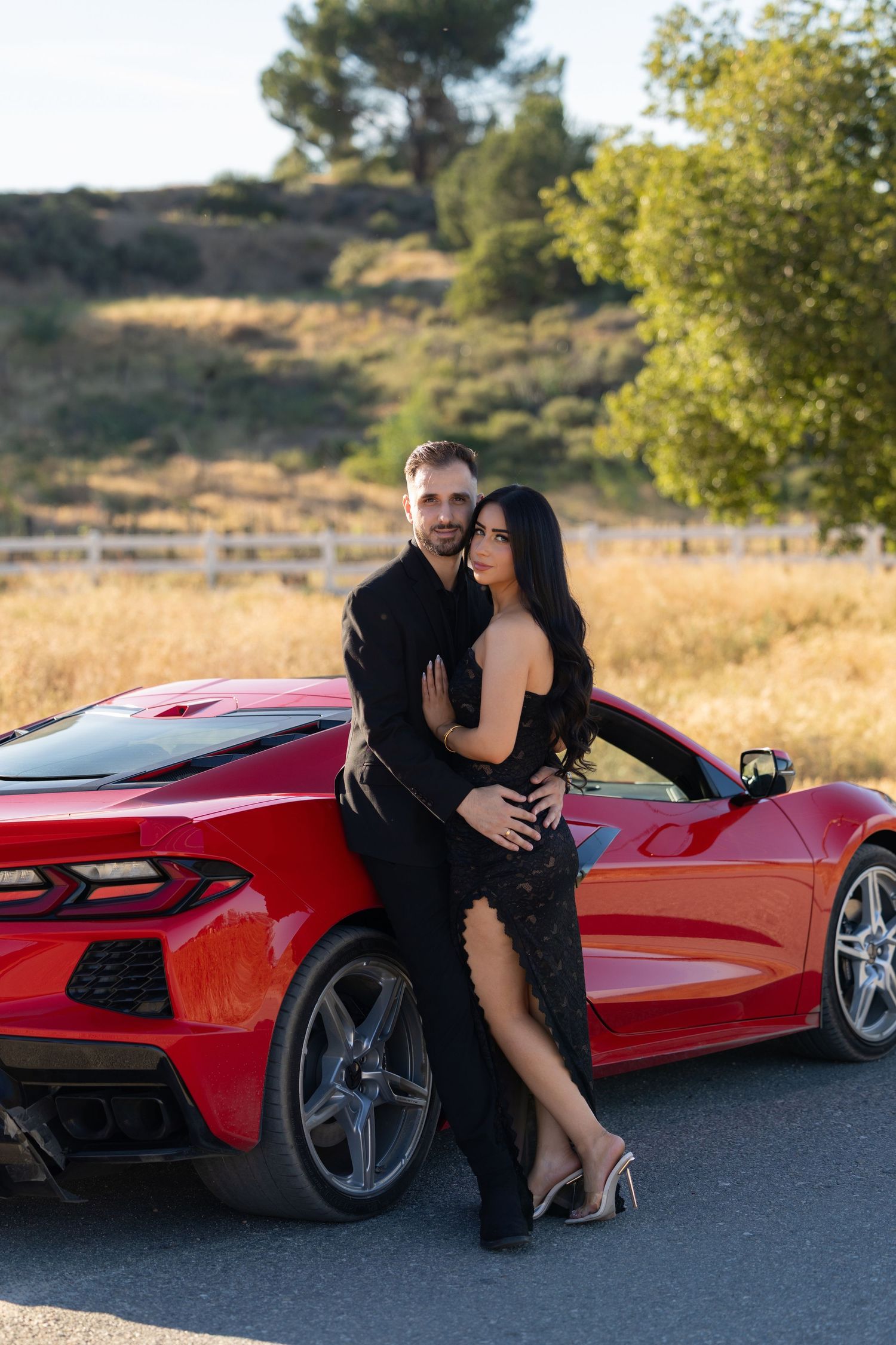 Couple in black formal wear posing romantically against a red Corvette sports car on a scenic road.
