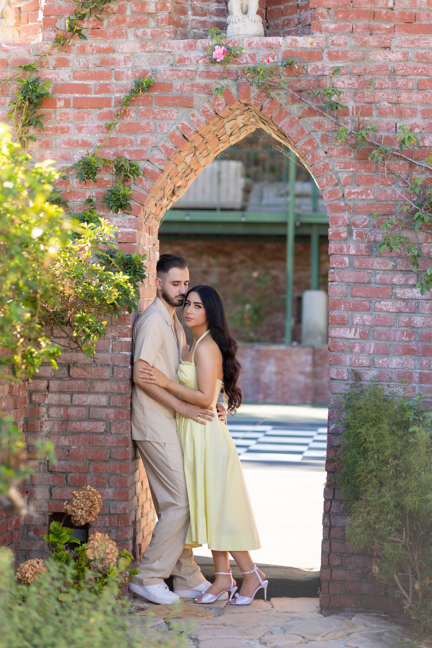 Couple embracing in a romantic brick archway surrounded by lush greenery, woman in yellow dress, man in beige outfit.