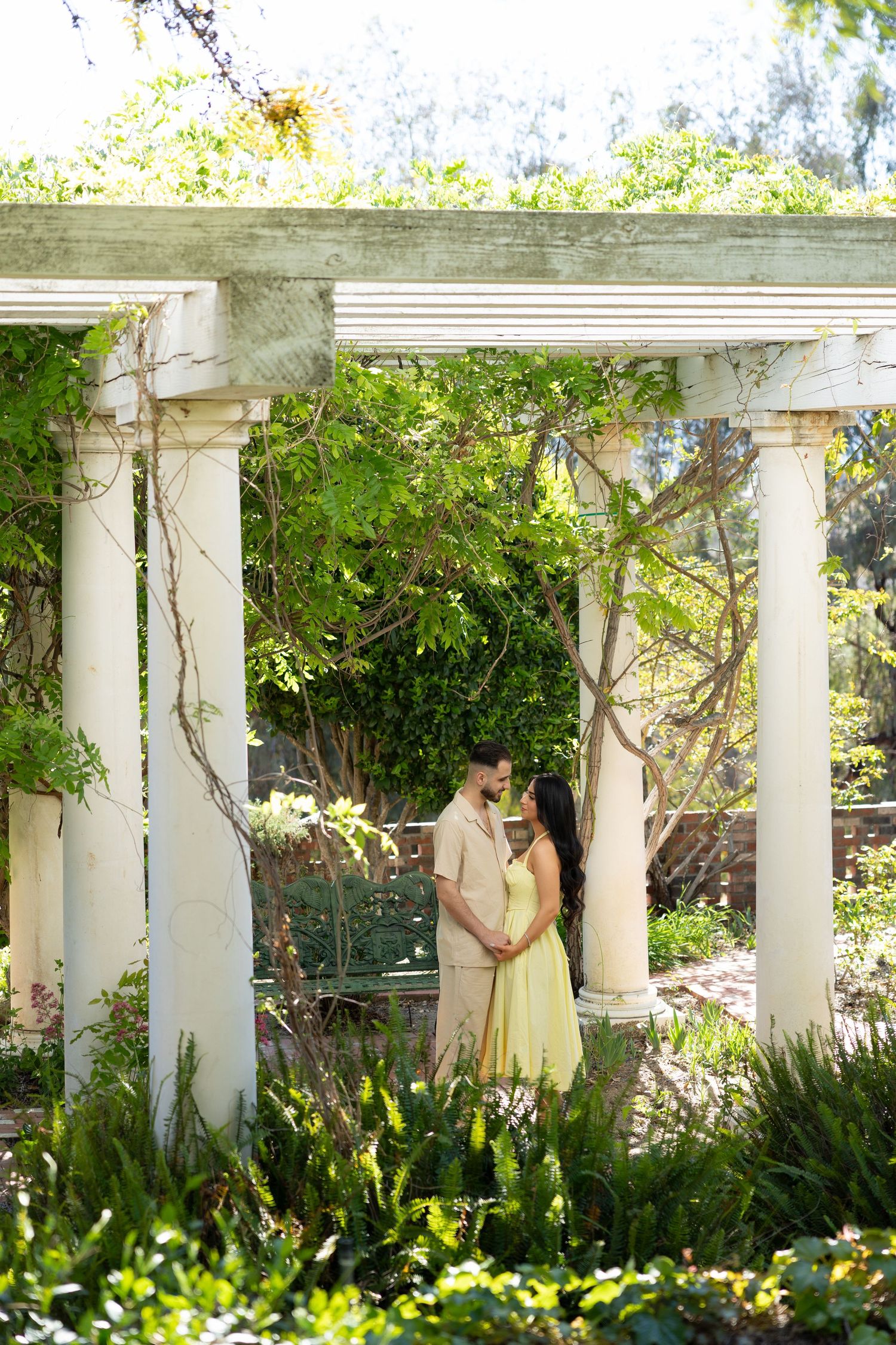 A couple embraces under a white columned pergola surrounded by lush green garden foliage in soft natural light.