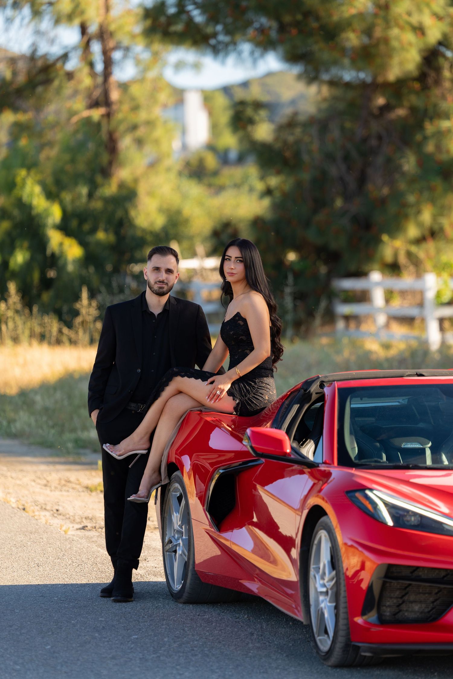 A couple in elegant black attire poses beside a red sports car on a scenic road surrounded by lush green trees.