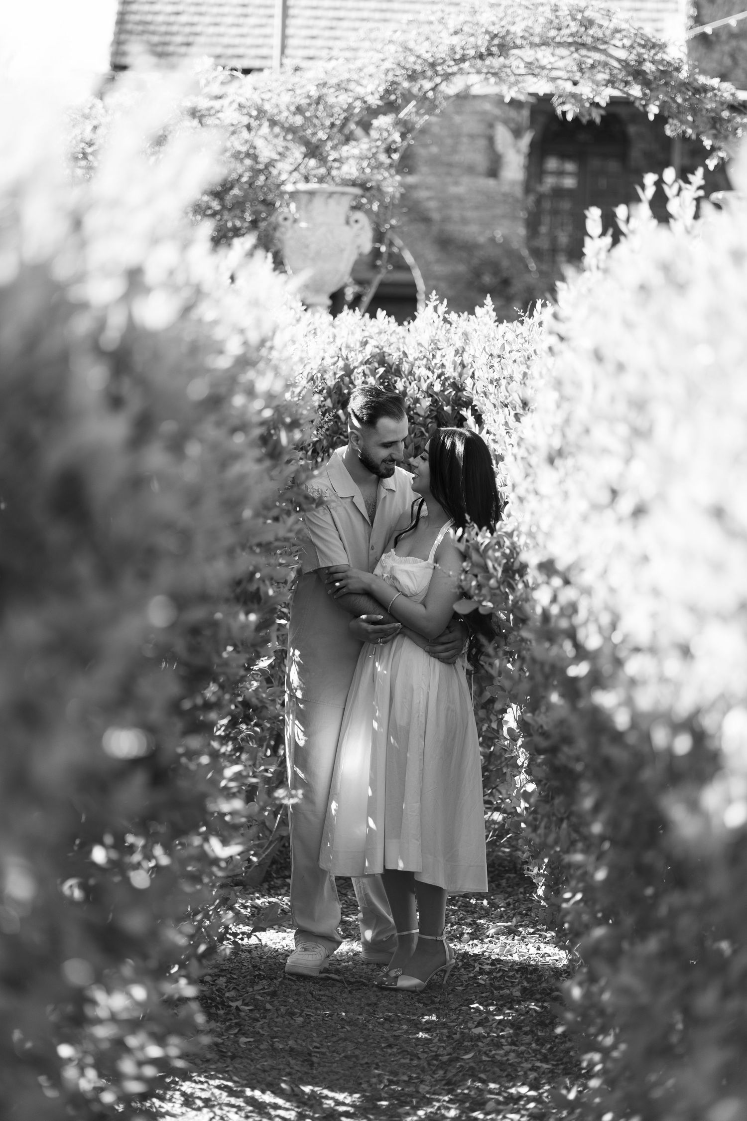 Black and white photo of a couple embracing in a lush garden pathway, surrounded by blooming flowers and greenery.