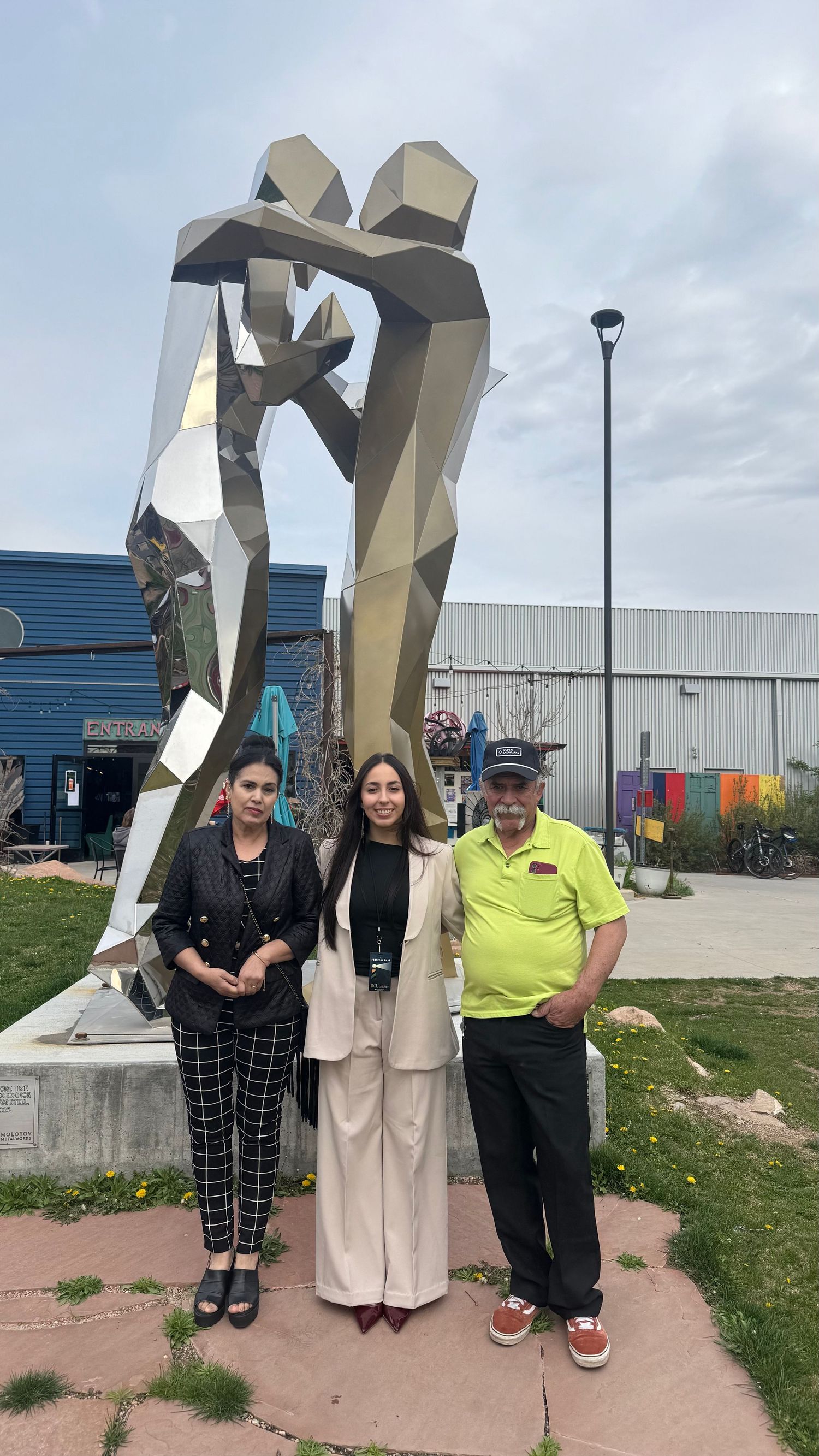 Three people posing in front of a large geometric metallic sculpture outdoors near colorful buildings.