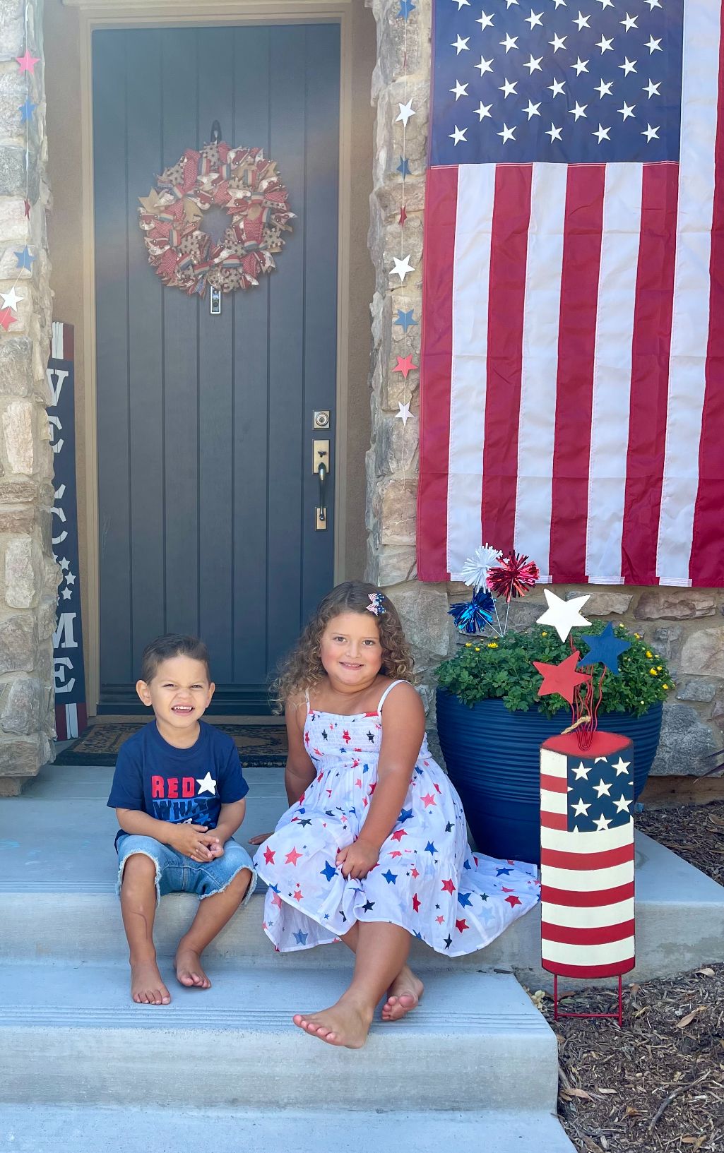 Patriotic front porch decor with American flag, floral wreath on blue door, and red white and blue planters on stone steps.