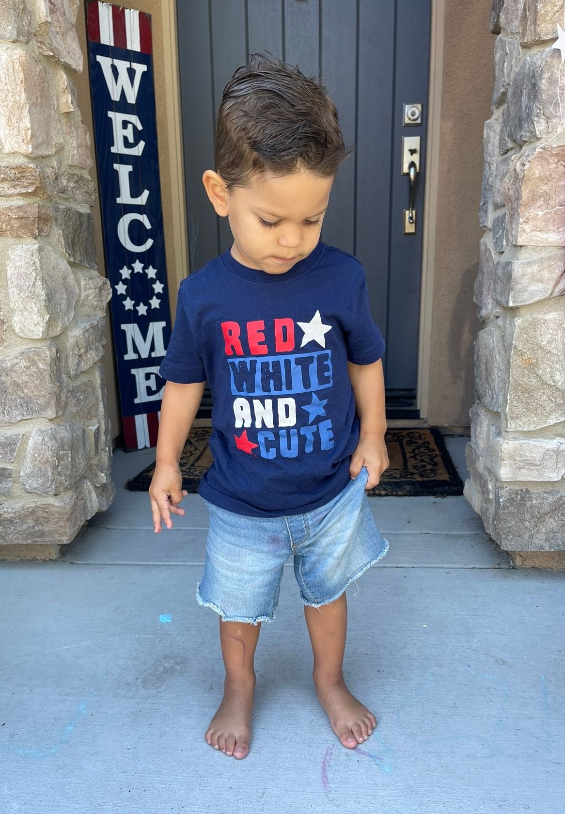 Child wearing patriotic red, white and blue t-shirt and denim shorts stands barefoot by welcome sign at front door.