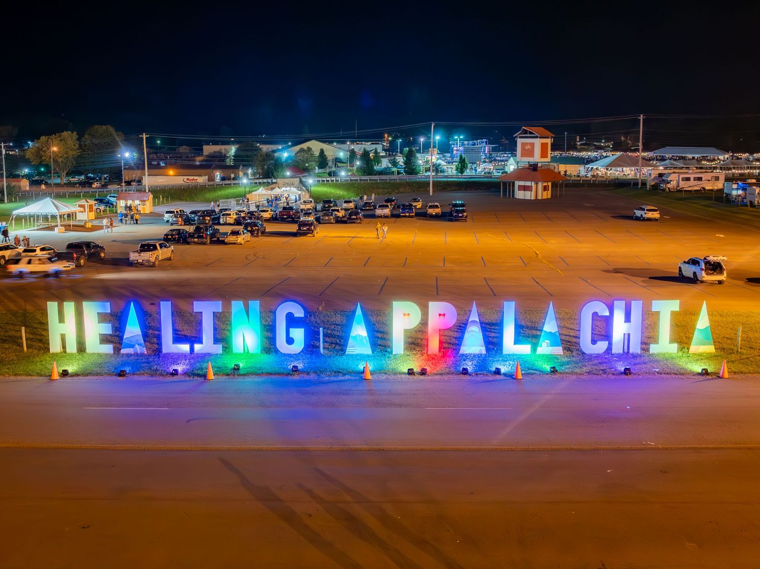 Colorful rainbow LED lights spell out 'HEALING APPALACHIA' at night in a parking lot with buildings visible in background.