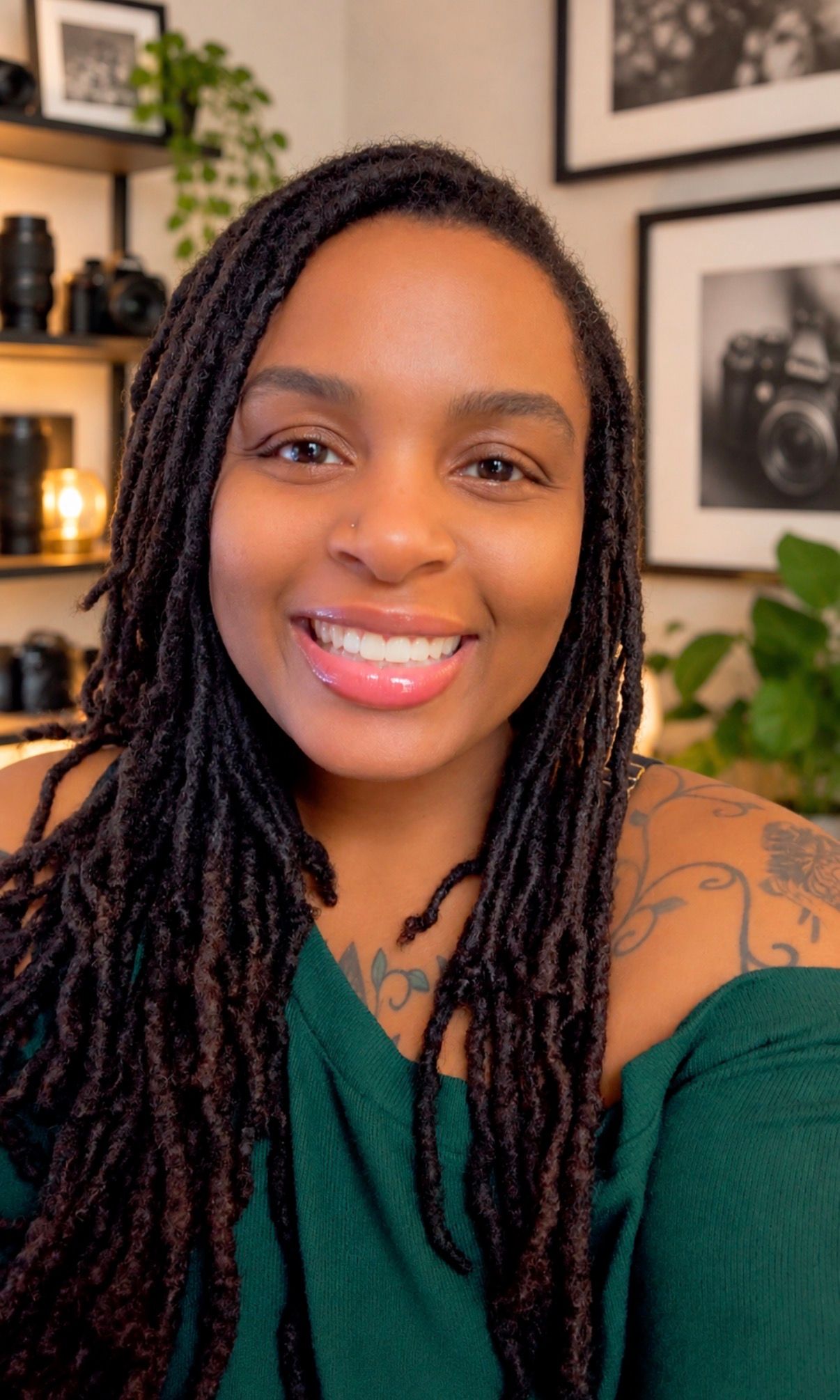 Smiling woman with long locs and shoulder tattoo poses in a green top in a photography studio.