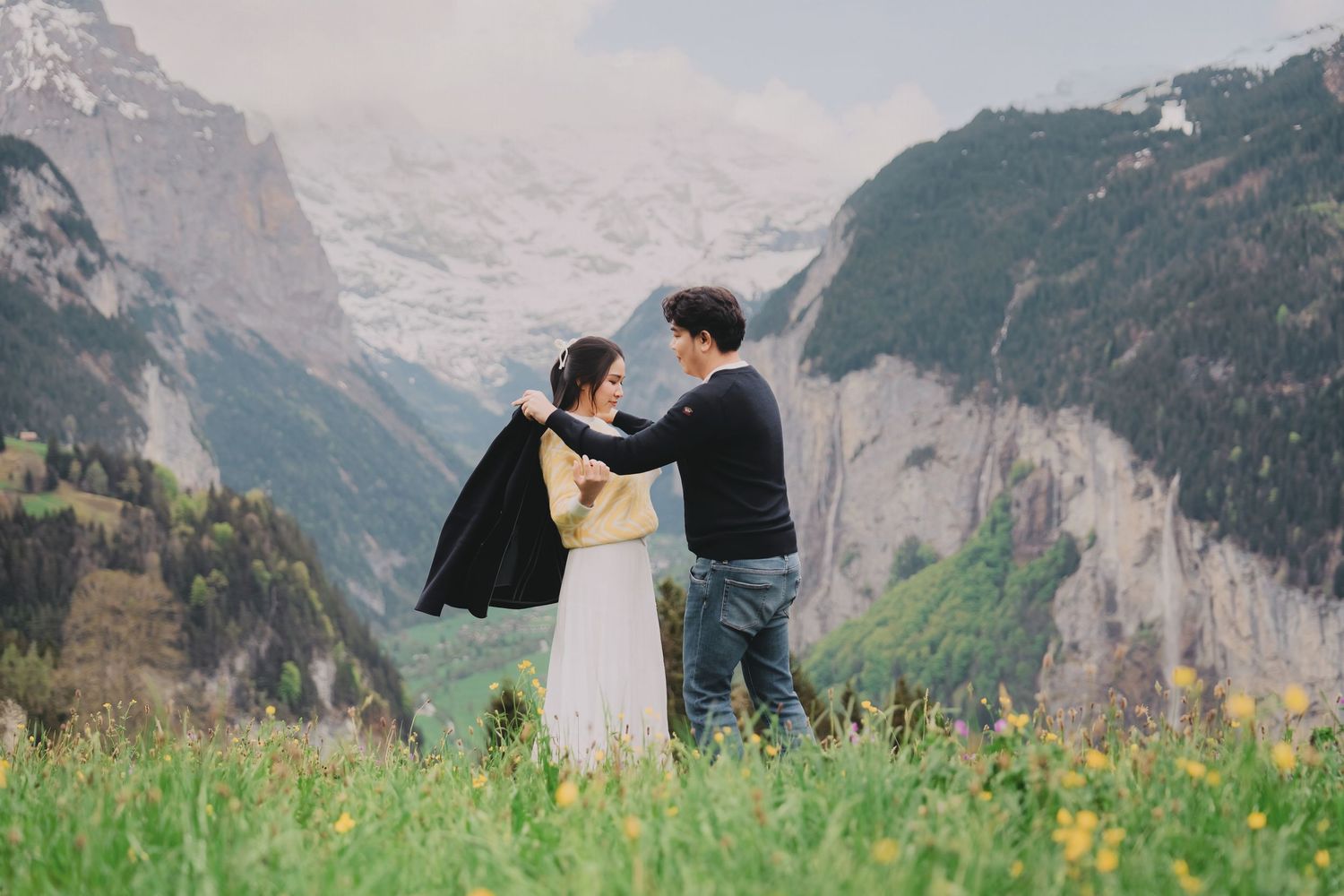 Man placing jacket on womans shoulders in flower meadow with Swiss Alps and waterfall behind.