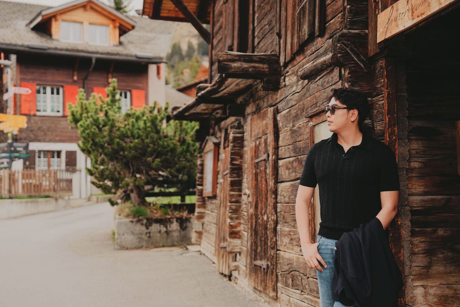 Stylish Asian man in black polo leaning against rustic wooden building wall in Swiss mountain village.