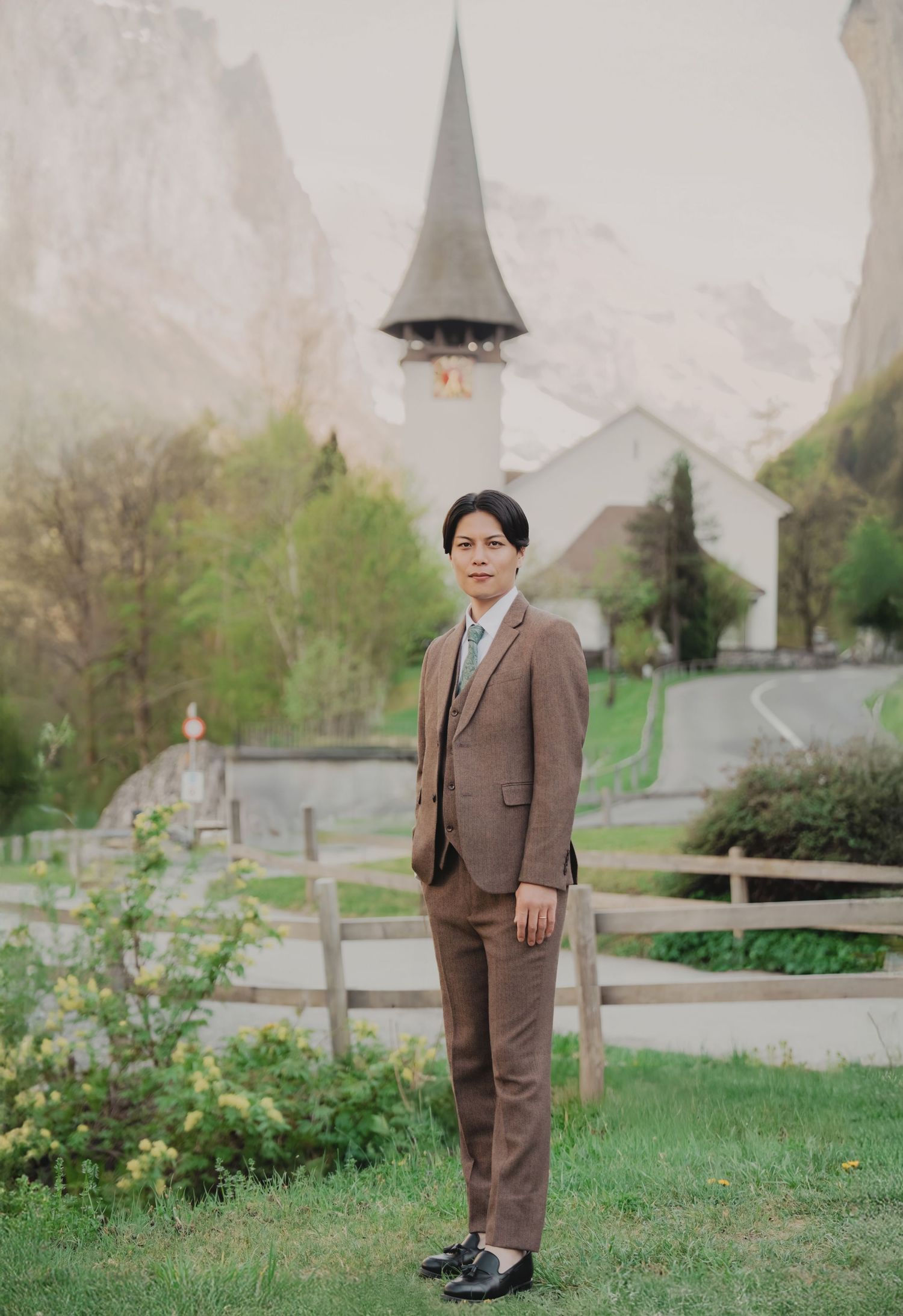 Groom in brown three-piece suit stands on green lawn with Swiss church backdrop.