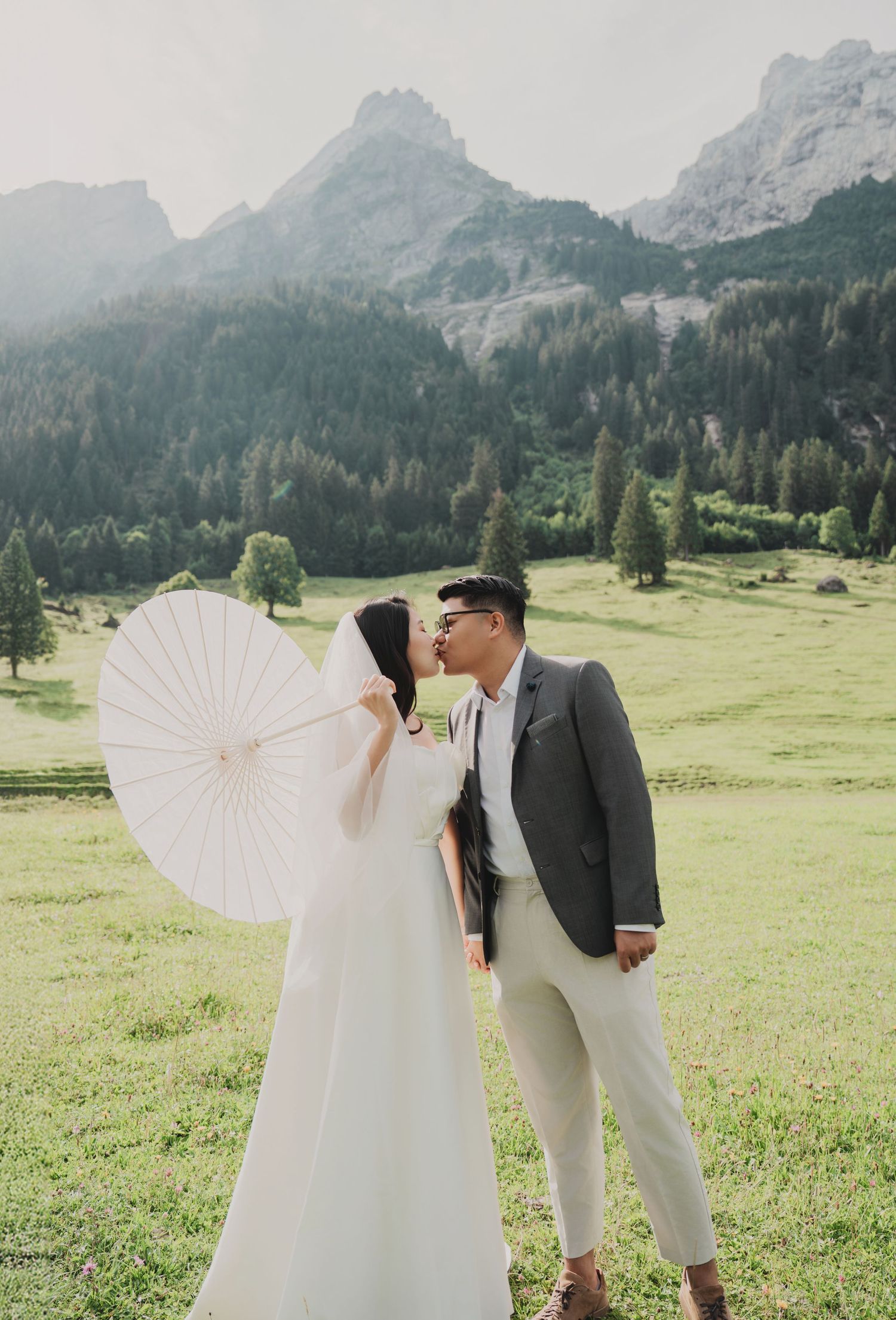 Couple kissing in Alpine meadow with white parasol and rocky mountain peaks in background.