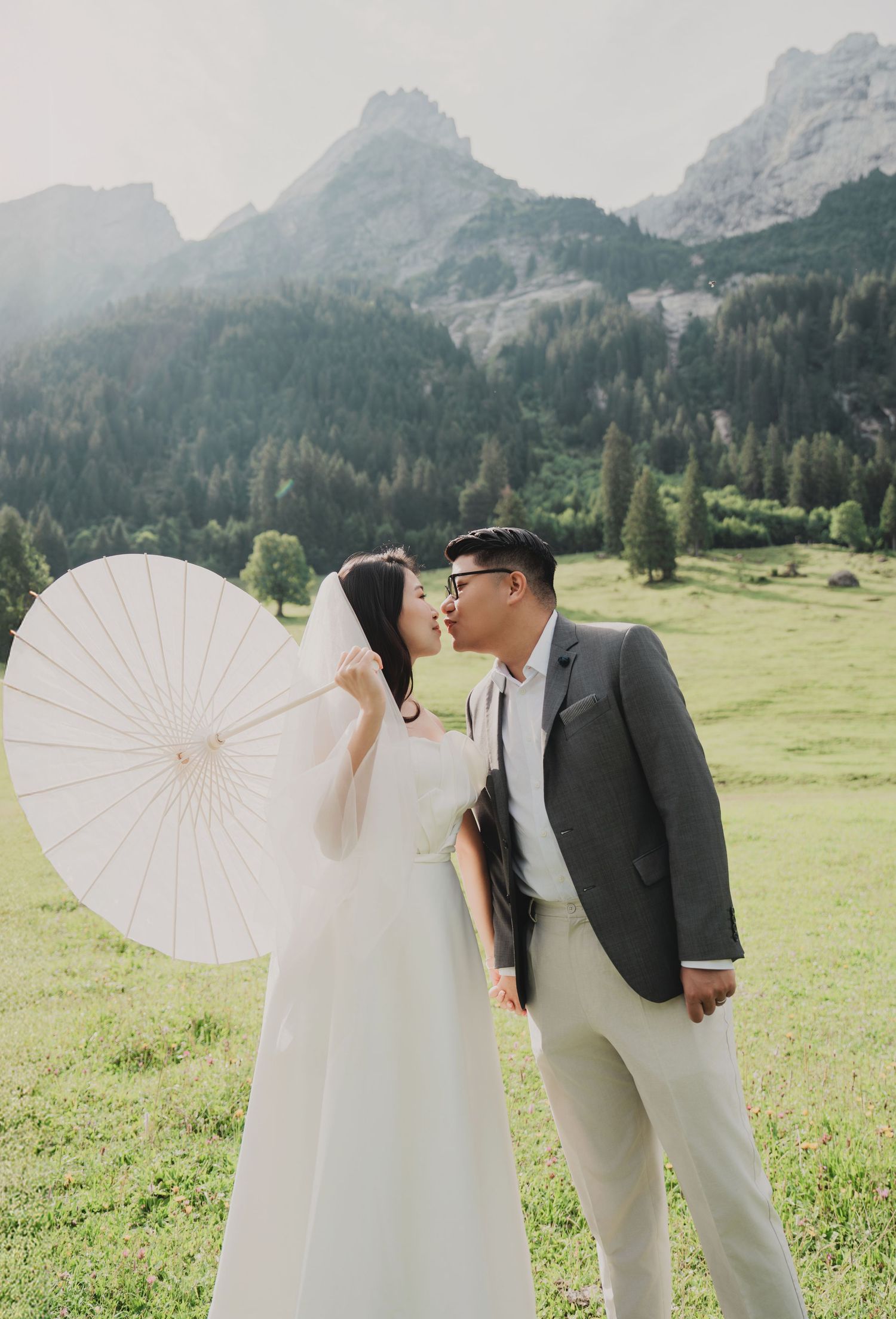 Asian couple shares a kiss in mountain meadow, bride in white gown holding decorative parasol with rocky peaks behind.