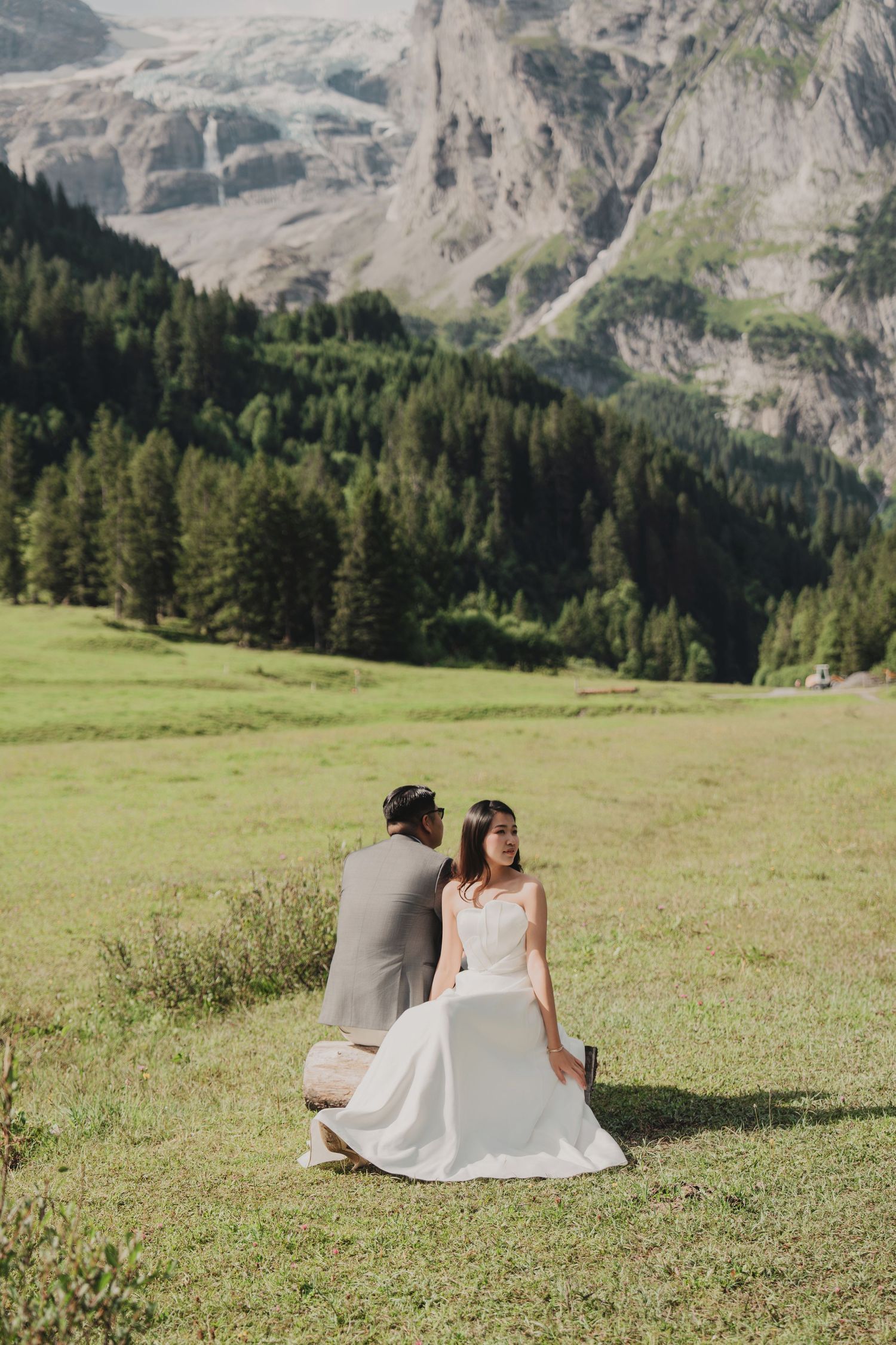 Bride and groom sitting on alpine meadow, bride gazing back with glacier mountain views behind.