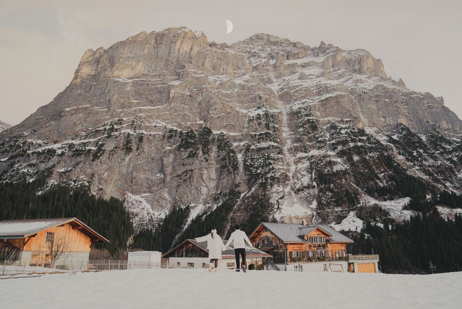 Snow-covered Alpine village with wooden chalets beneath a massive rocky mountain and crescent moon.