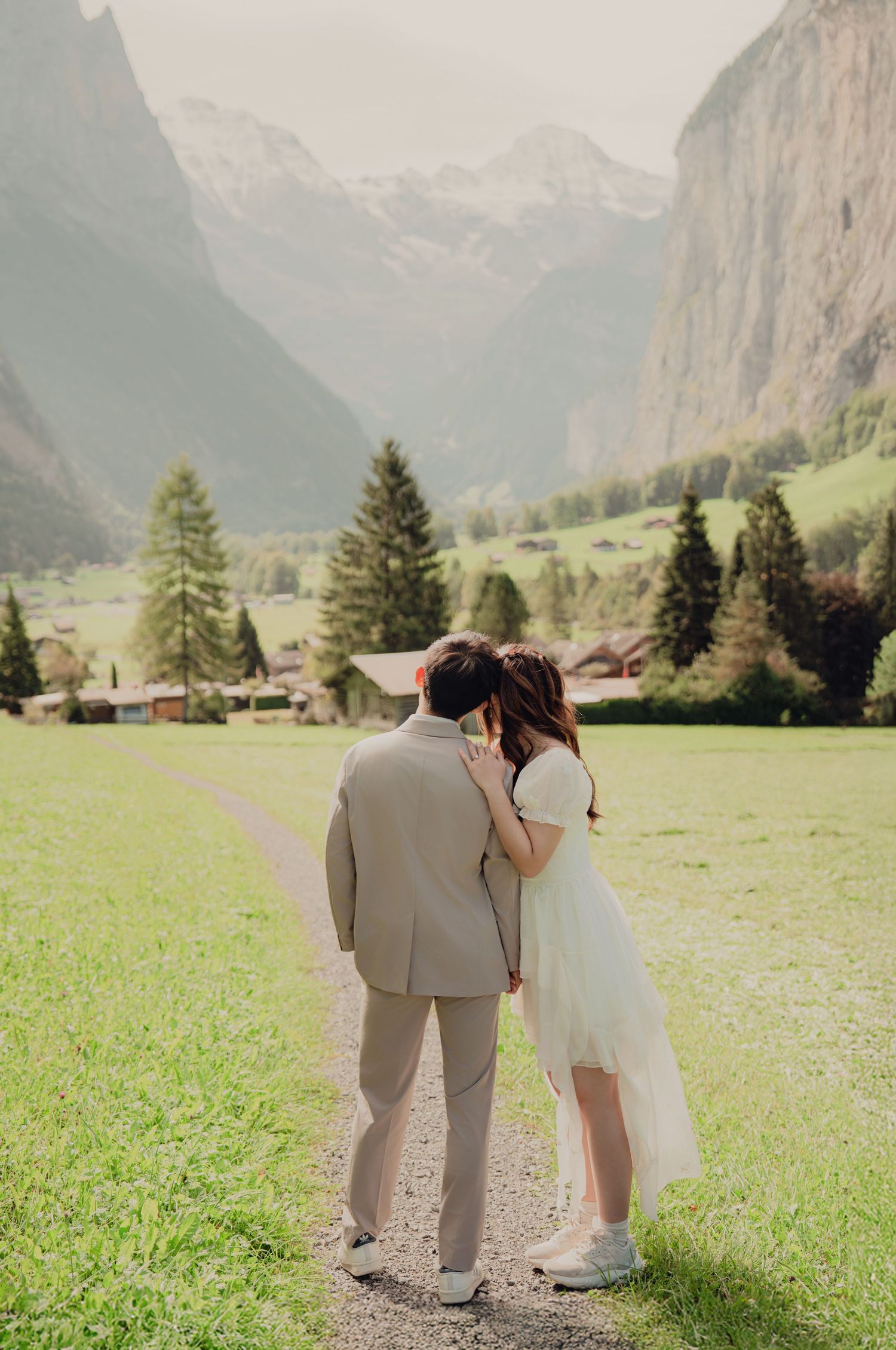 Wedding couple embracing on a grassy path with stunning Swiss Alpine valley and mountains in the background.
