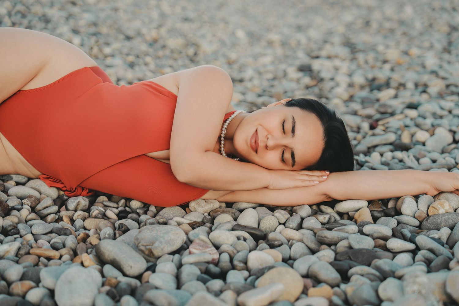 Woman in red swimsuit lying on pebble beach with eyes closed, smiling peacefully.