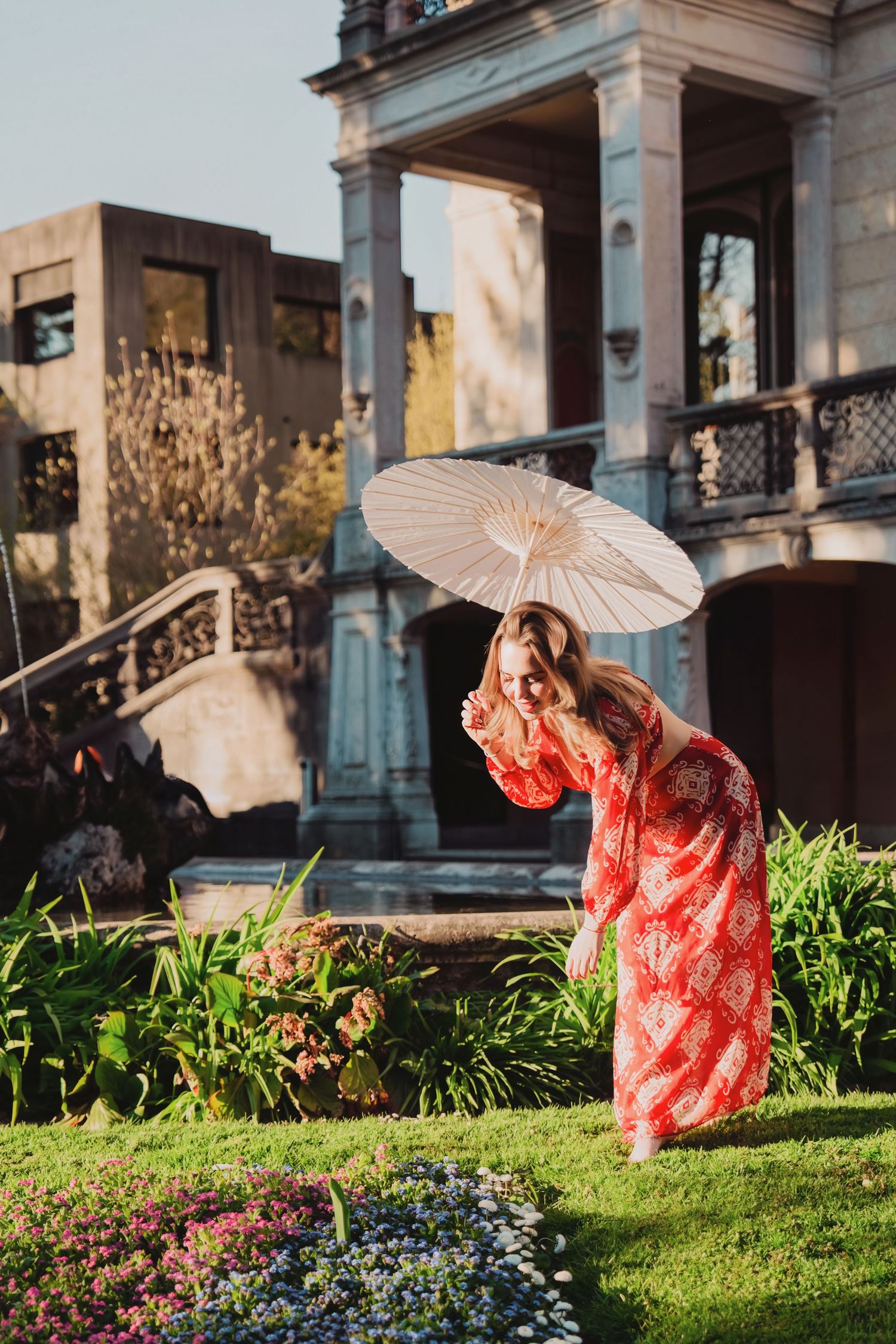 Woman in red floral dress with parasol in garden near historic building.