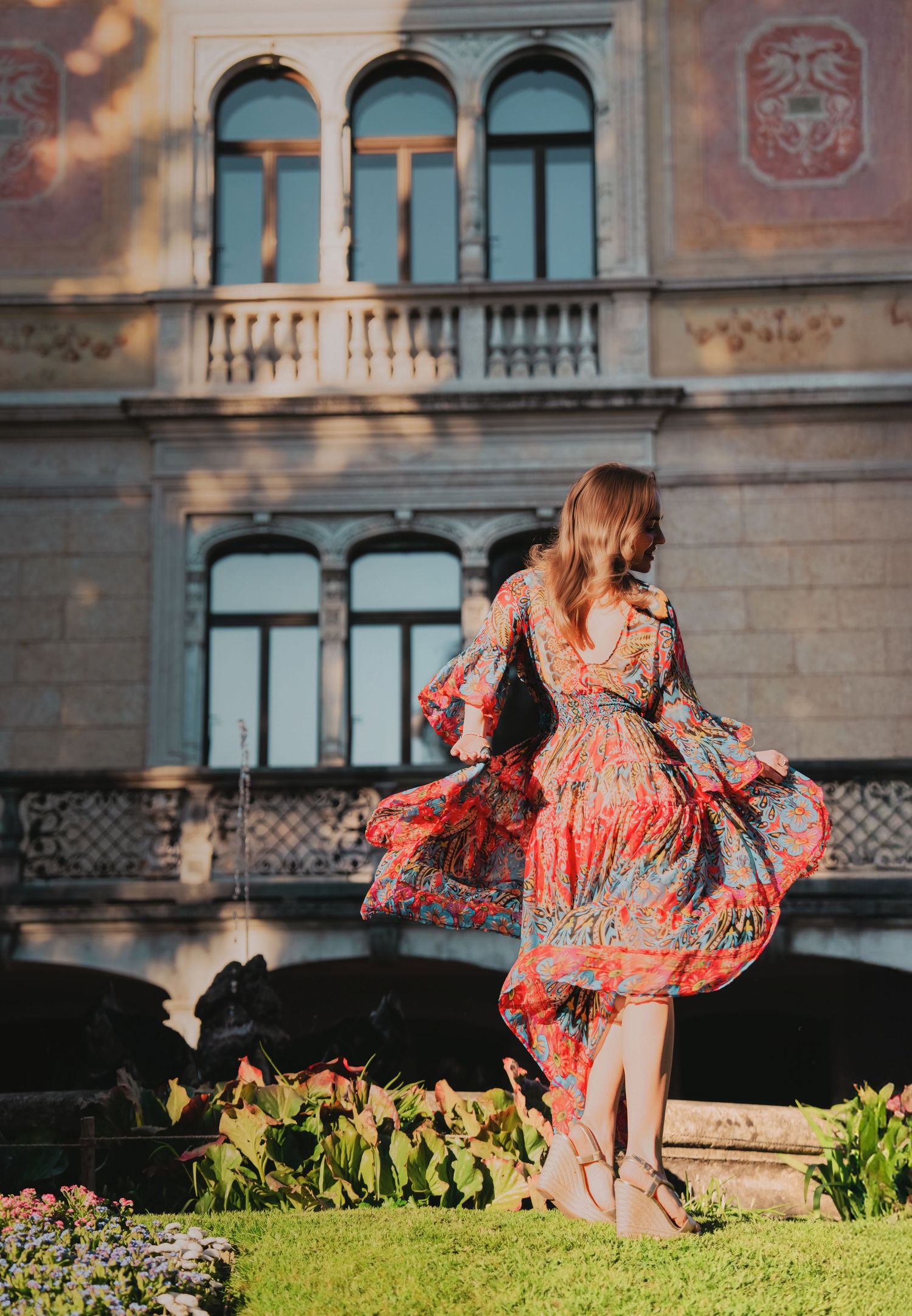 Woman in colorful floral dress twirling in golden sunlight before ornate European building facade.