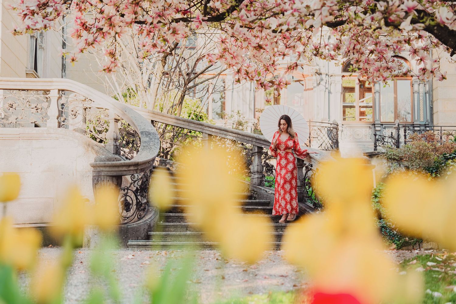Woman in red floral dress on ornate steps surrounded by blooming cherry blossoms and yellow tulips in spring garden.