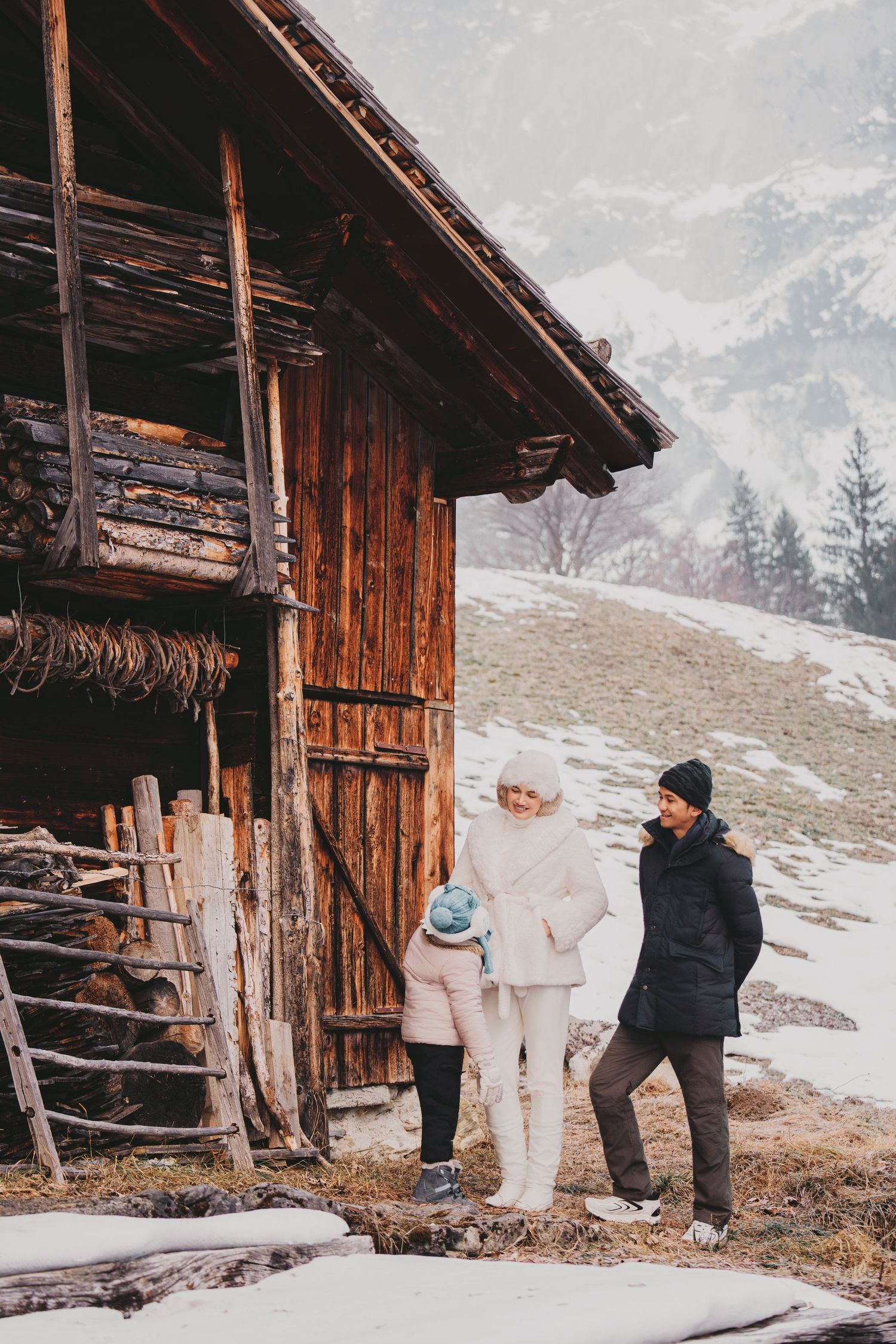 Family of three in winter coats standing beside rustic wooden barn with snowy mountains behind.