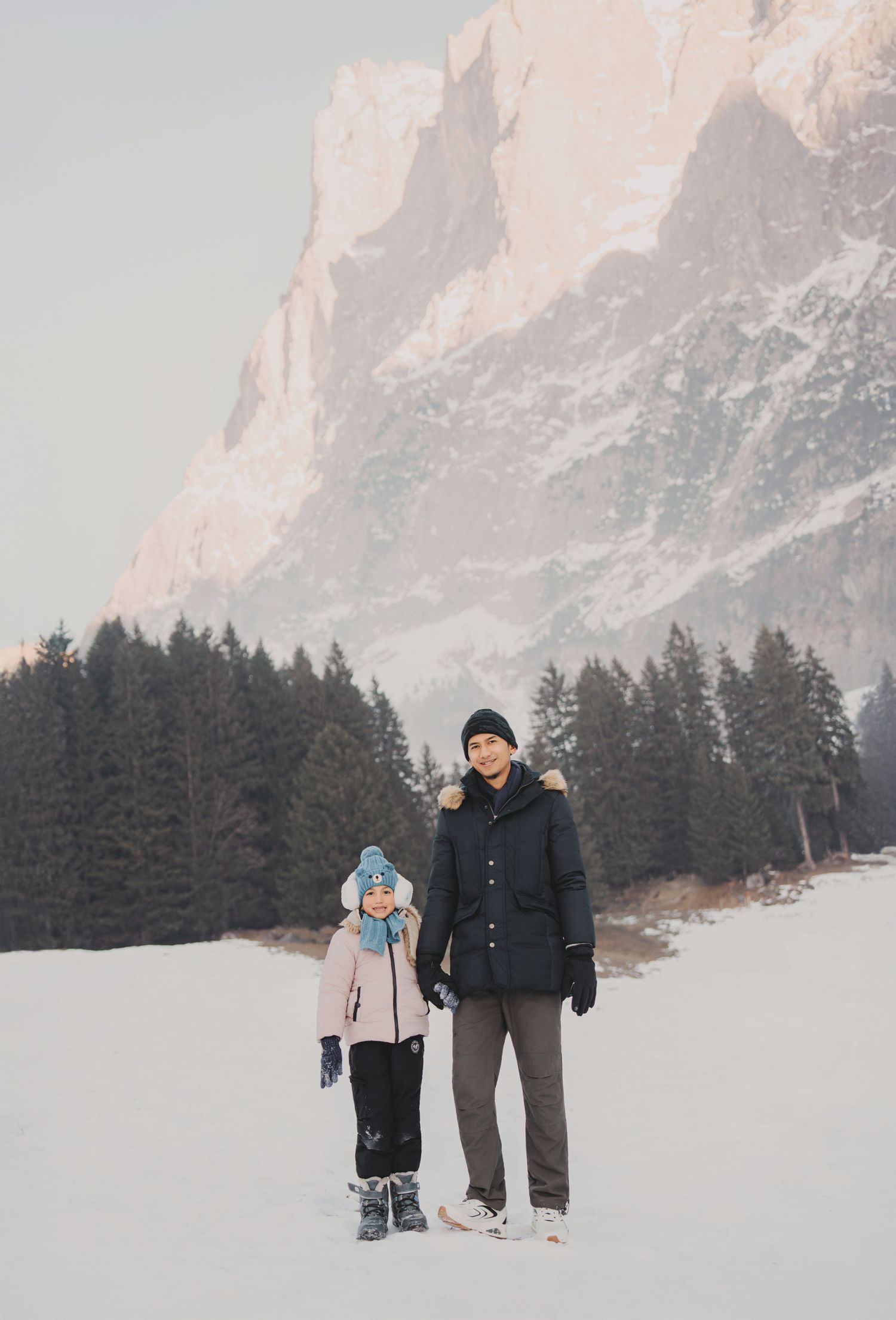 Man and child standing together in snowy field with tall rocky mountains and pine trees behind them.