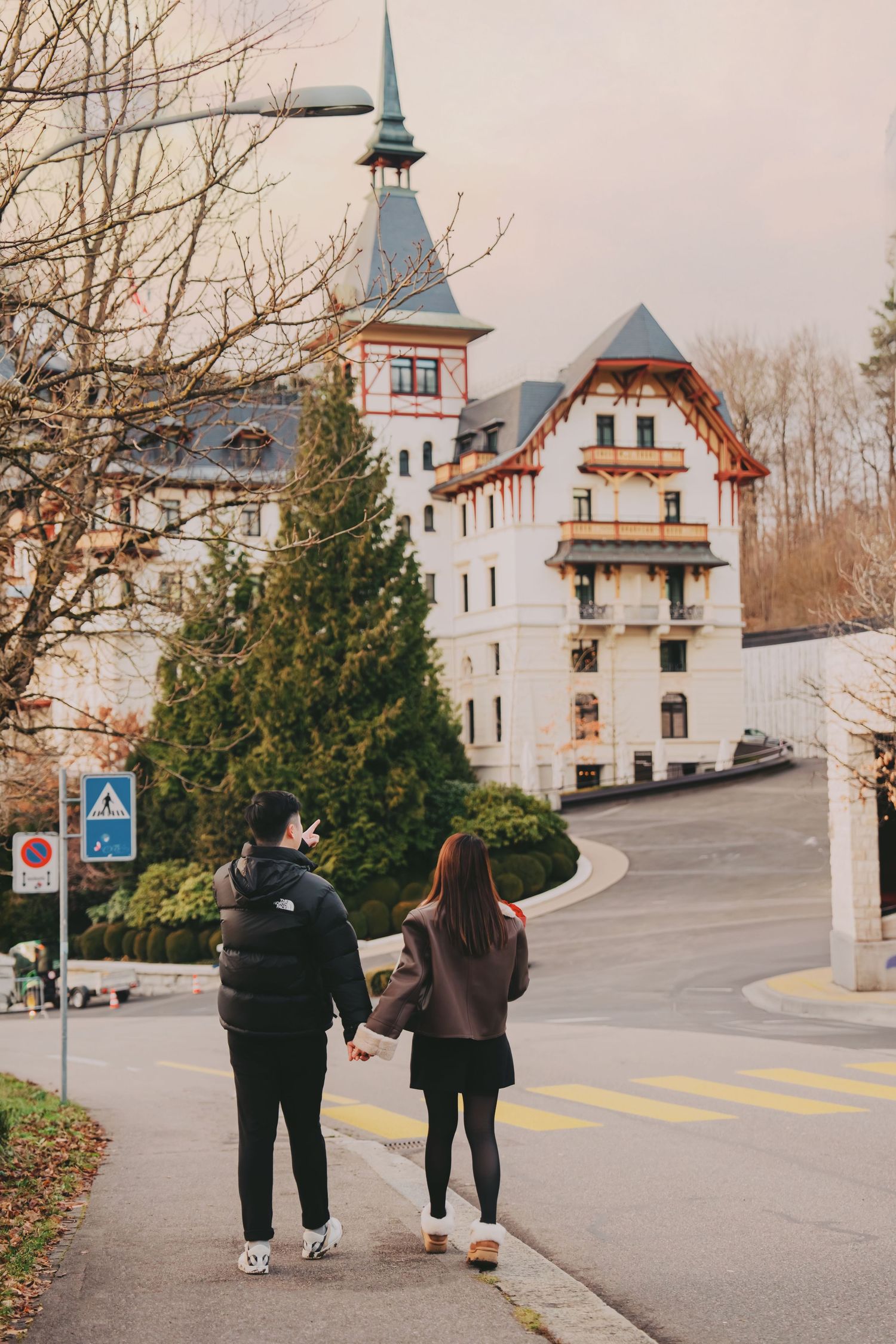 Couple walking hand in hand along road toward charming European villa with green tree.